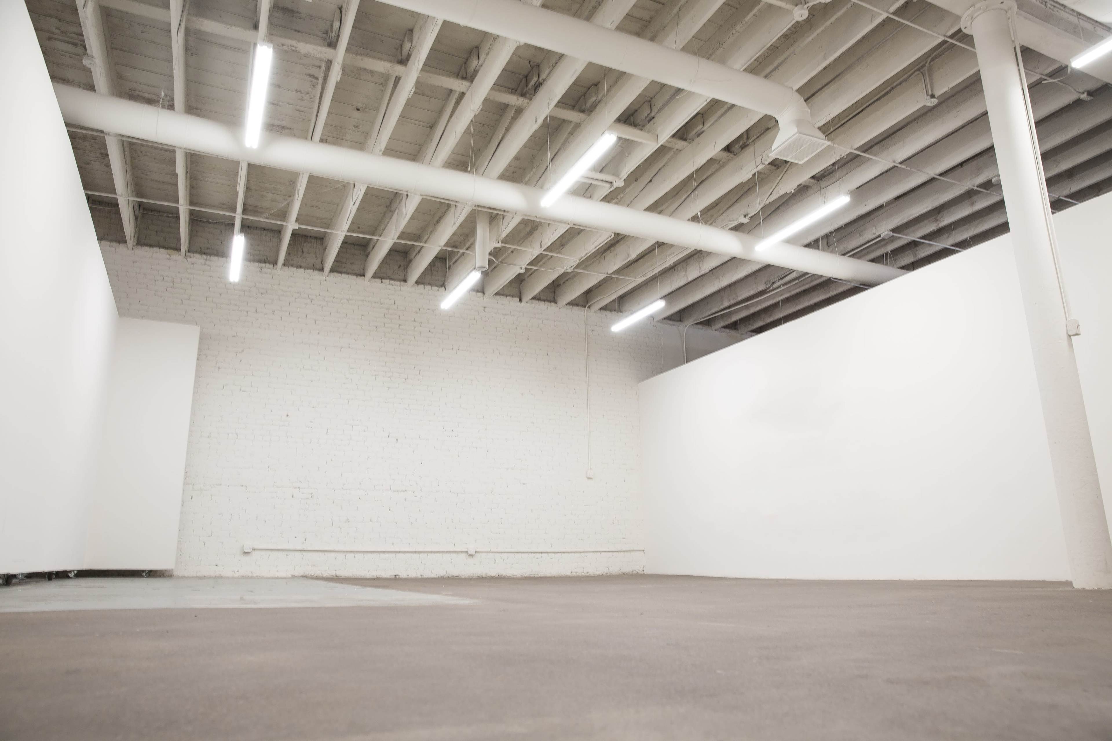 A spacious, empty room with white walls and a concrete floor, featuring industrial lighting overhead.