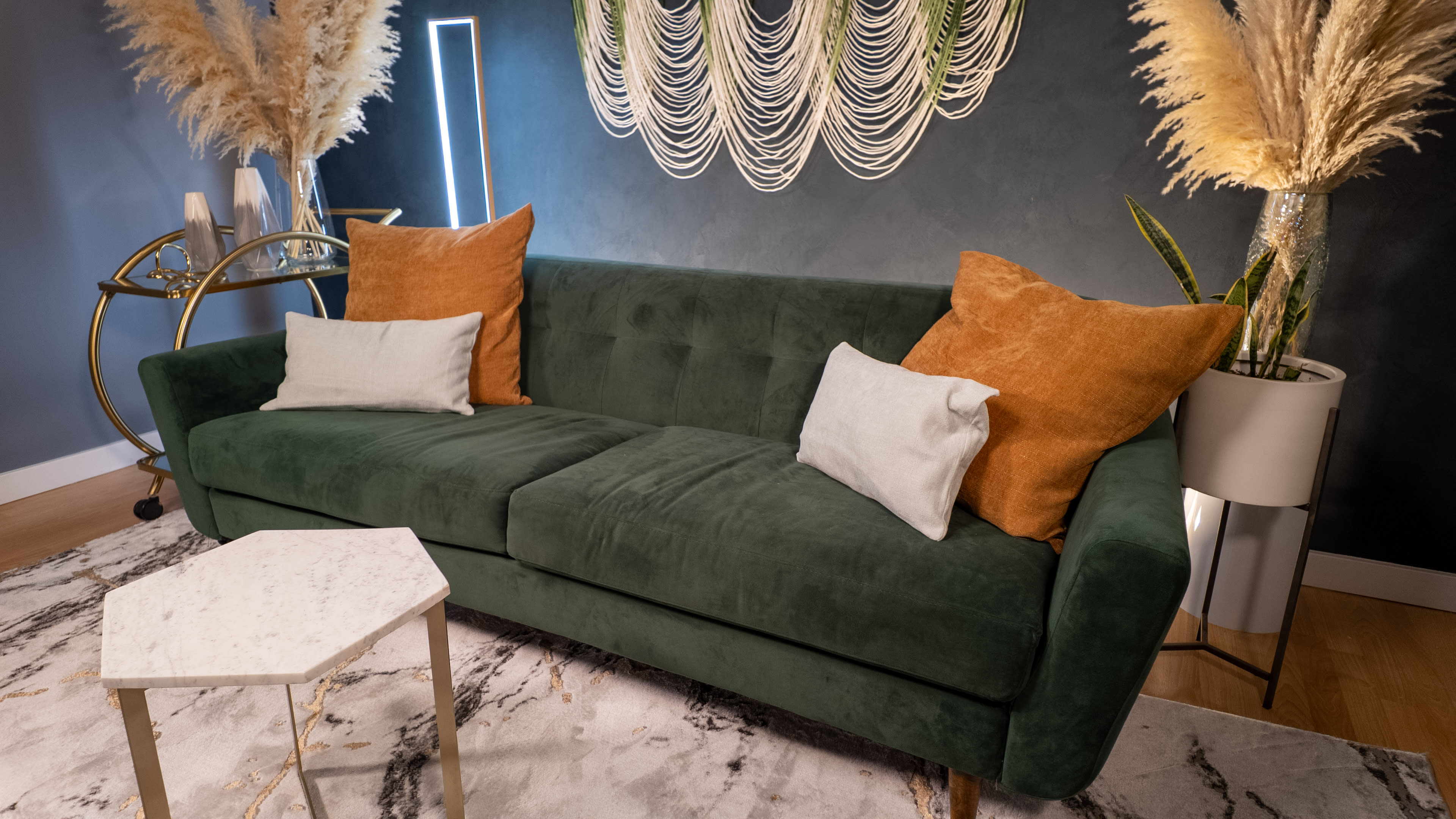 A green velvet sofa with orange and white pillows is positioned in a room featuring a marble-patterned rug, decorative plants, and wall decor.