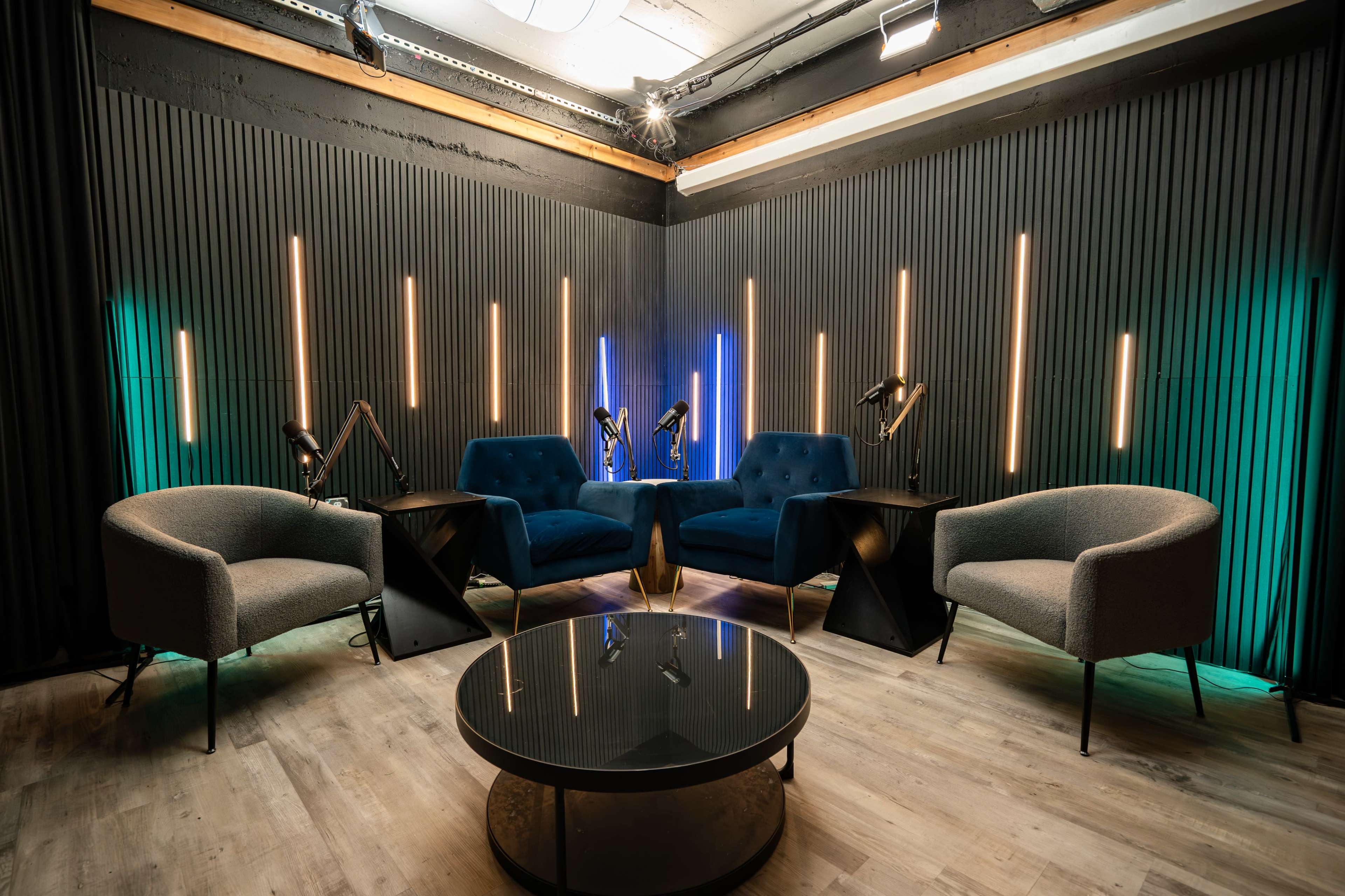The image features a modern seating area with two blue tufted armchairs, two gray chairs, and a round black coffee table, all set against a backdrop of vertical wooden slats illuminated by colorful LED lights.