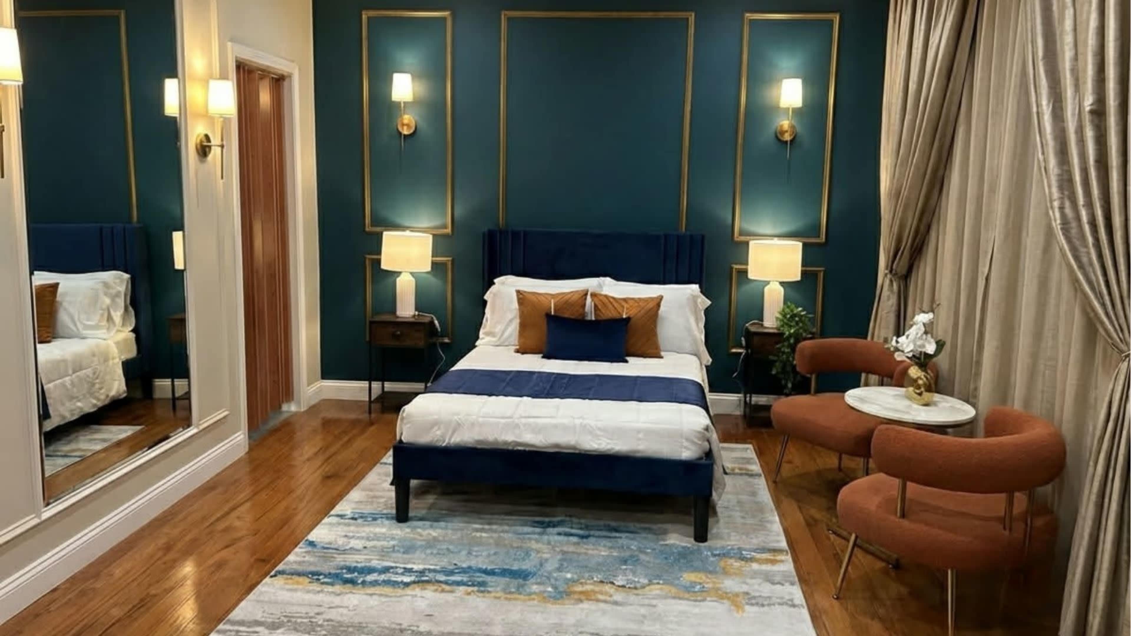 The image shows a stylish bedroom featuring a blue and white bed, two round armchairs, a large mirror, and decorative wall panels.