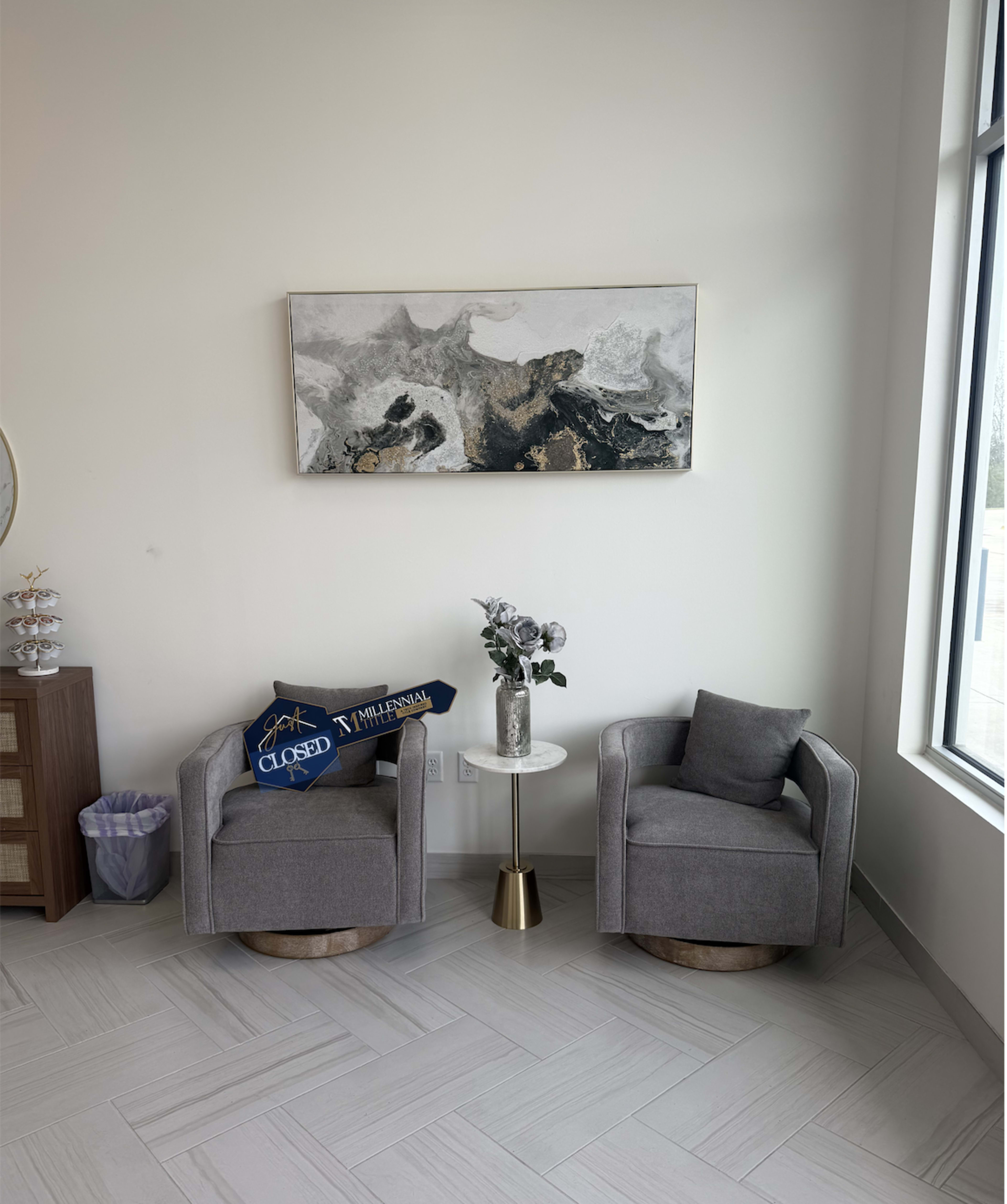 The image shows a bright, modern seating area with two gray swivel chairs, a small white side table holding a vase with flowers, and a wall-mounted abstract painting.