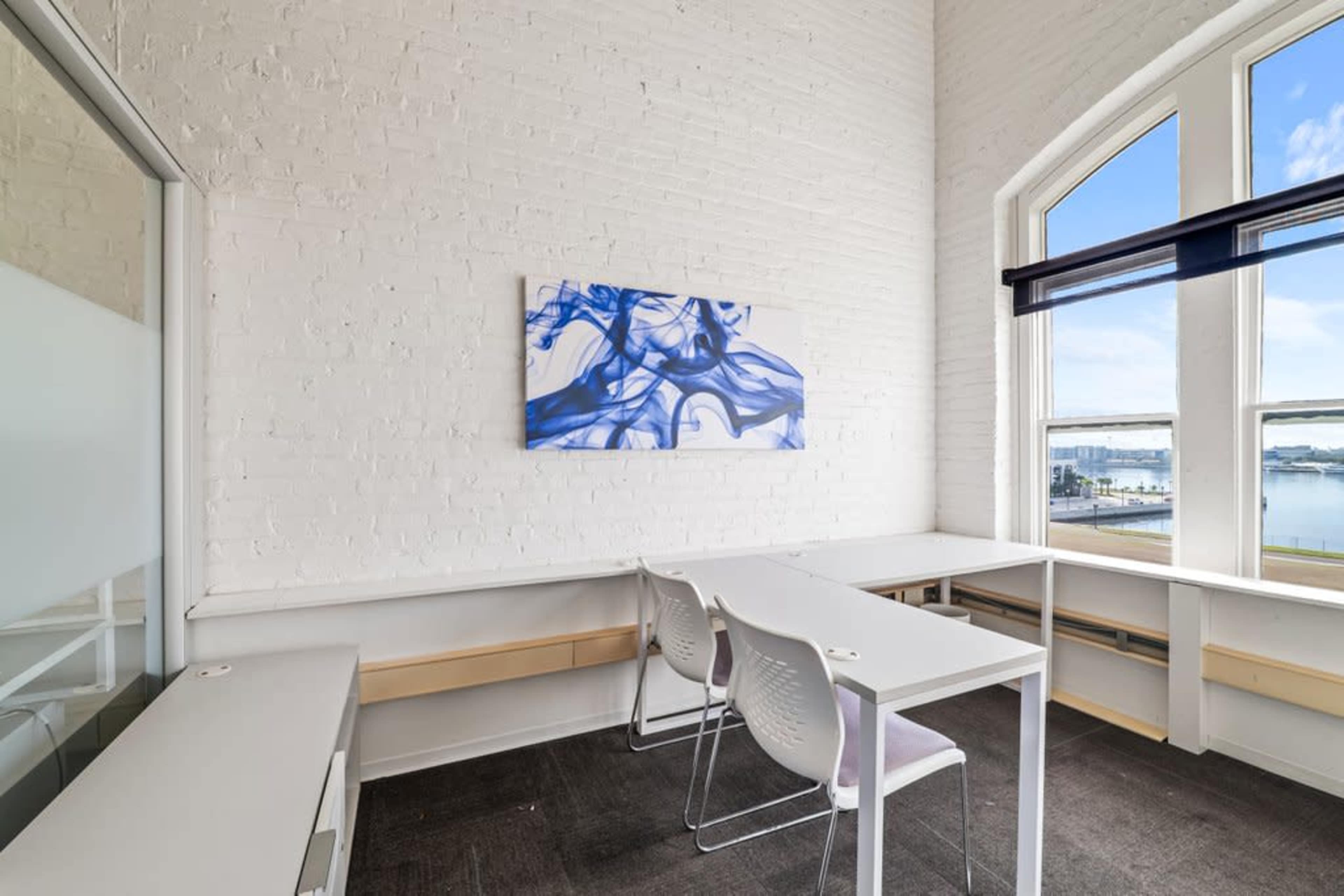 A bright, modern workspace features a large window, two white chairs, a minimalist desk, and a piece of abstract blue artwork on the wall.