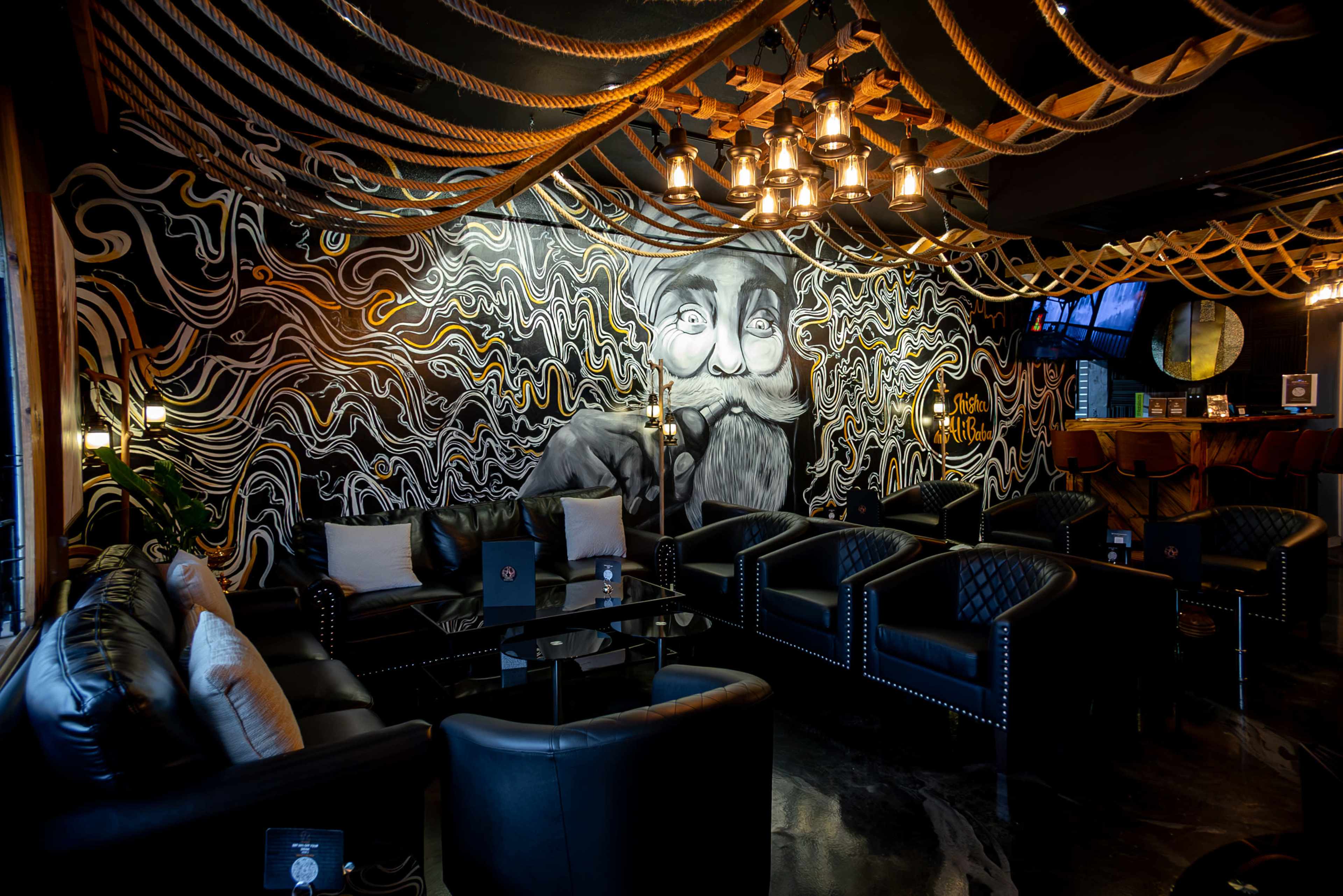A cozy lounge with black leather seating arranged around a dimly lit space featuring a vibrant mural and decorative ropes overhead.