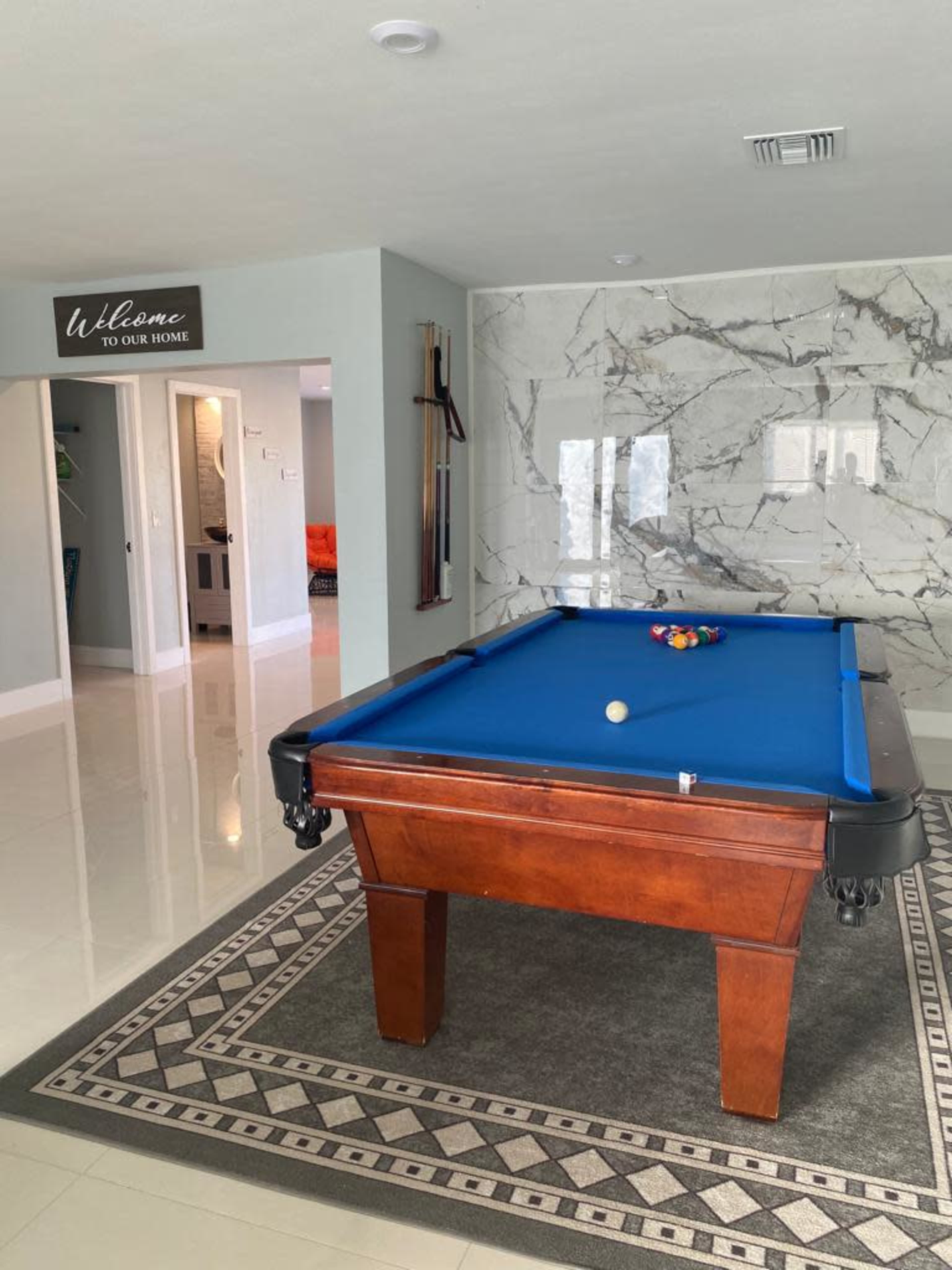 The image shows a brightly lit room featuring a blue pool table on a patterned rug, with a marble wall and an entrance leading to another part of the house.