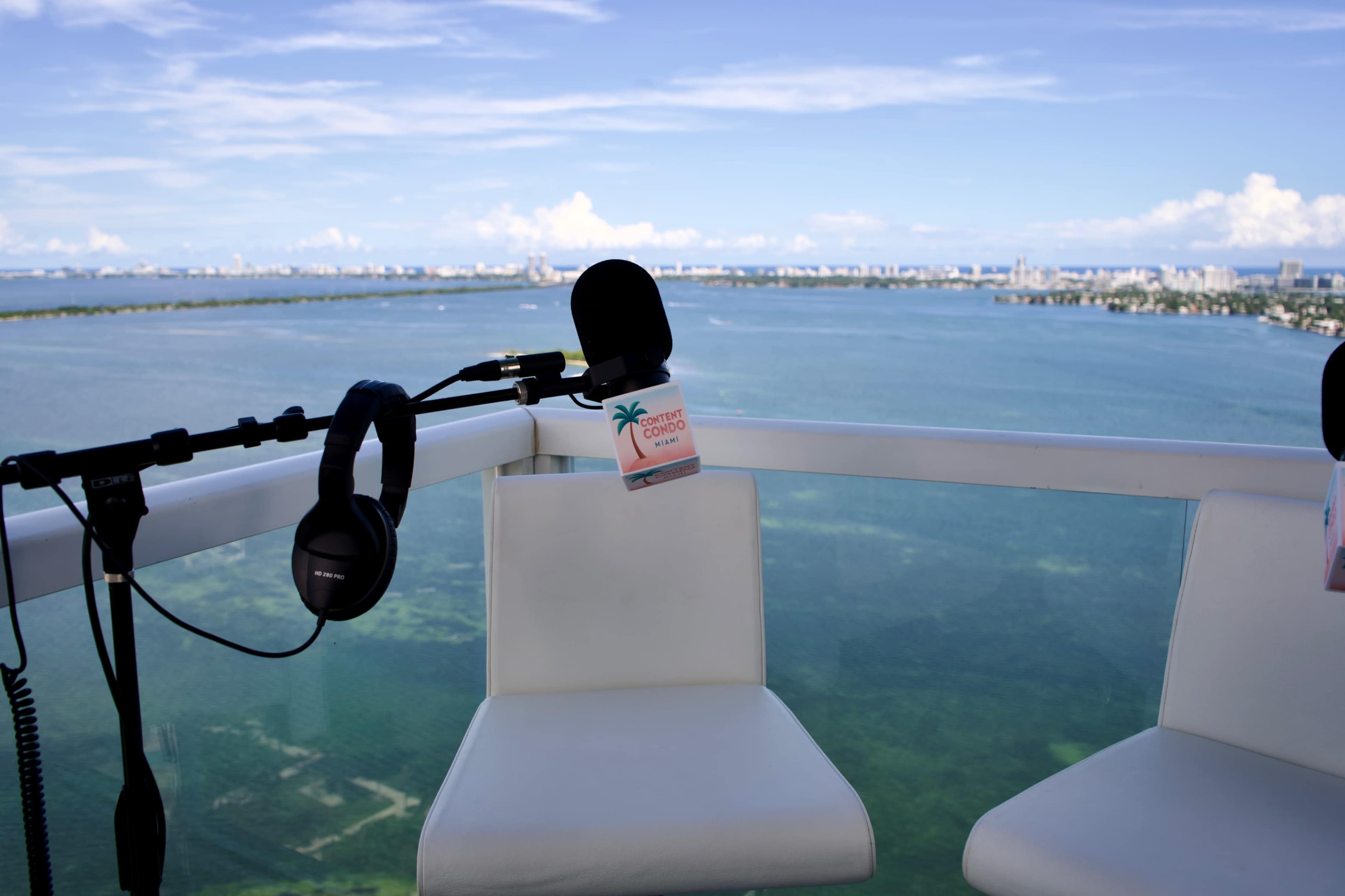 The image shows a microphone and headphones on a balcony overlooking a body of water with a city skyline in the distance.