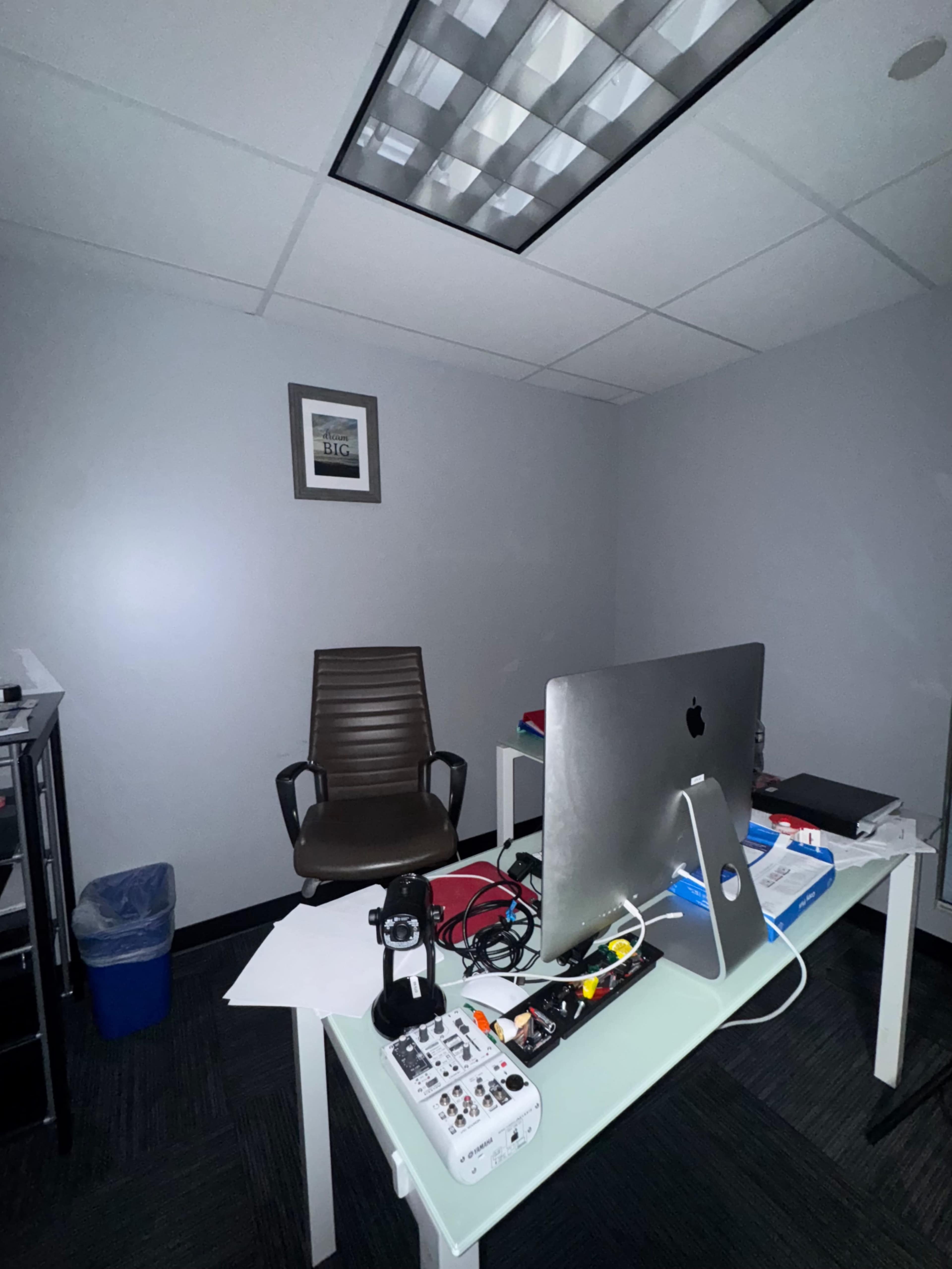 The image depicts a small office space featuring a desk with an iMac, an office chair, and various office supplies scattered on the desk.