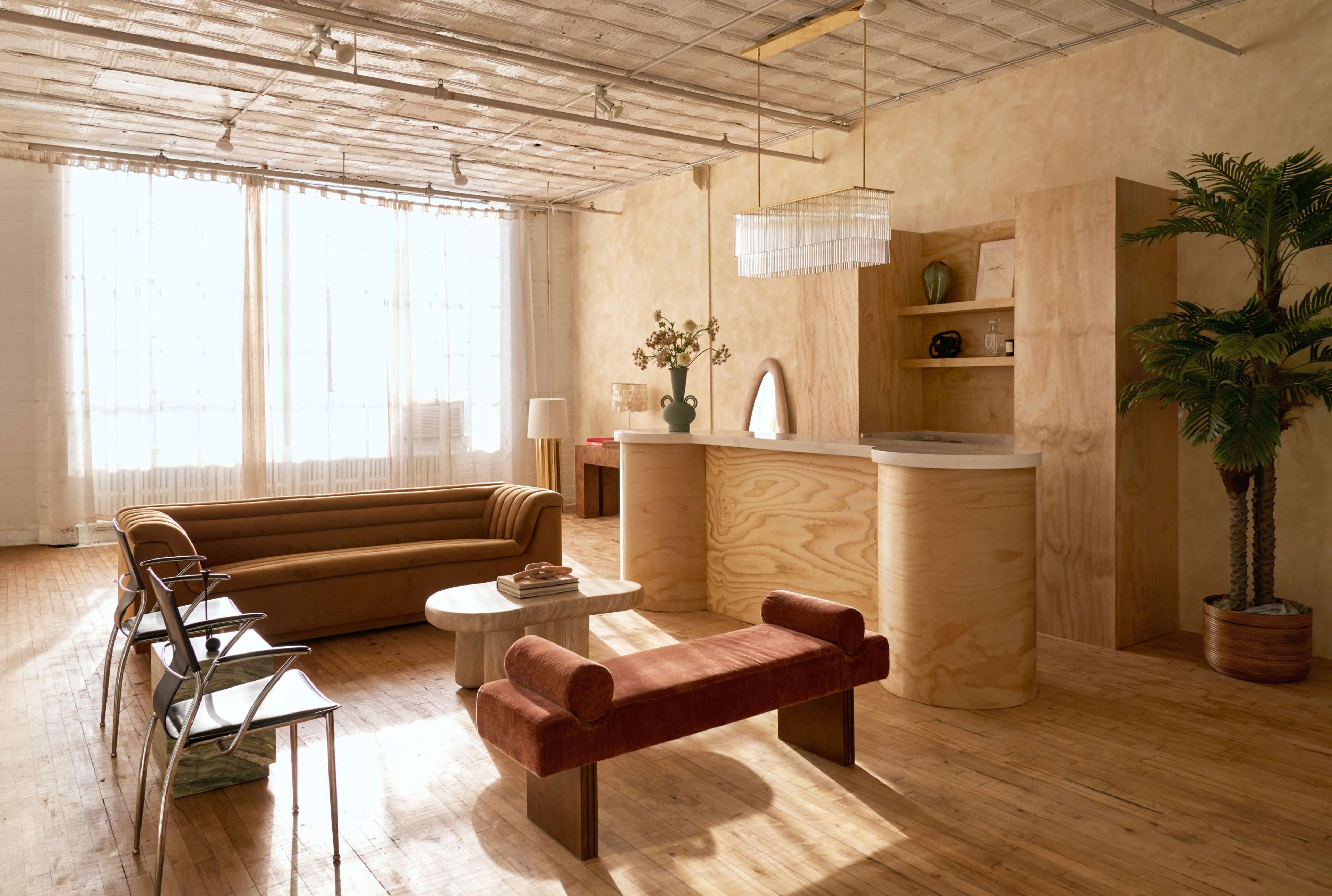 The image shows a spacious, well-lit interior featuring a brown sofa, a wooden reception counter, a circular coffee table, and potted plants.