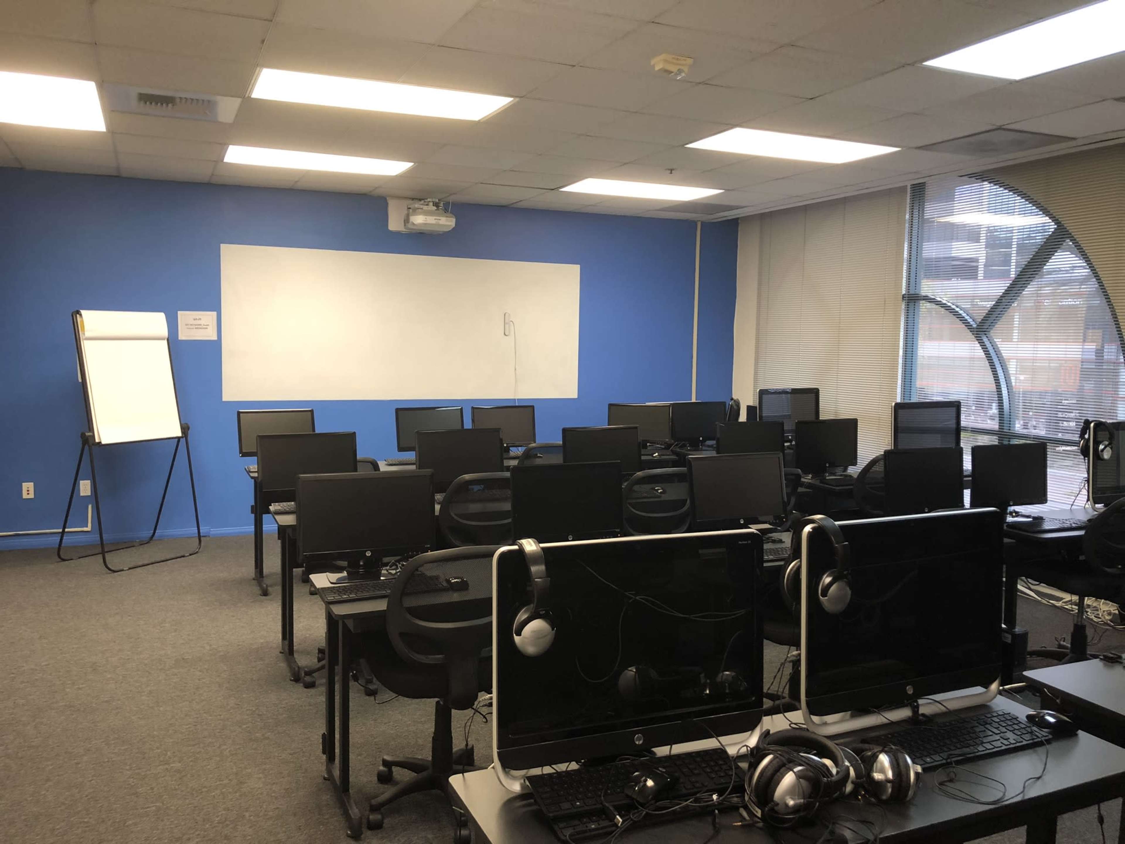 The image shows a computer lab with multiple desks equipped with monitors and headsets, and a whiteboard on the blue wall.