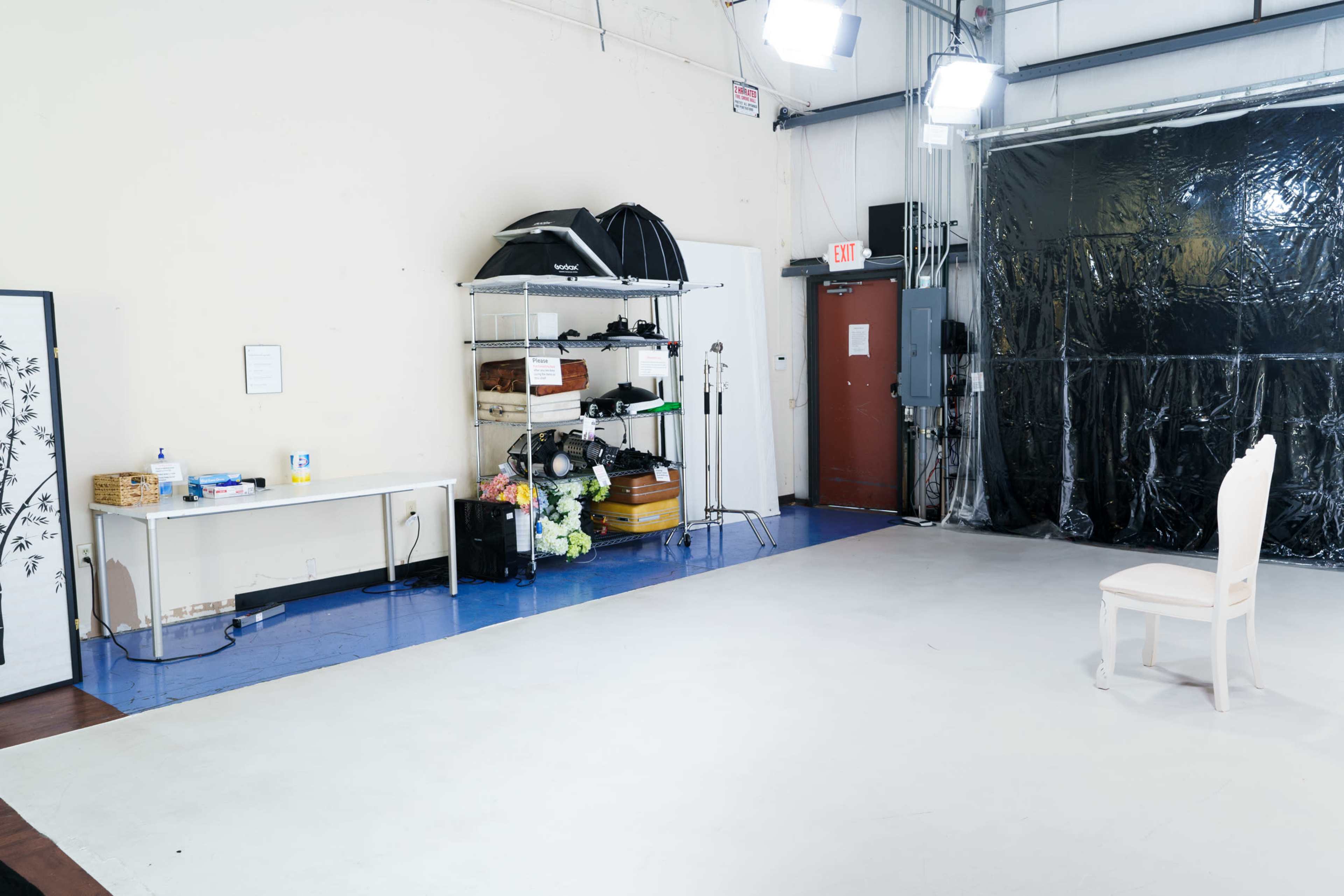 A spacious studio with a white floor, a single chair, storage shelves filled with various items, and black drapes covering one wall.
