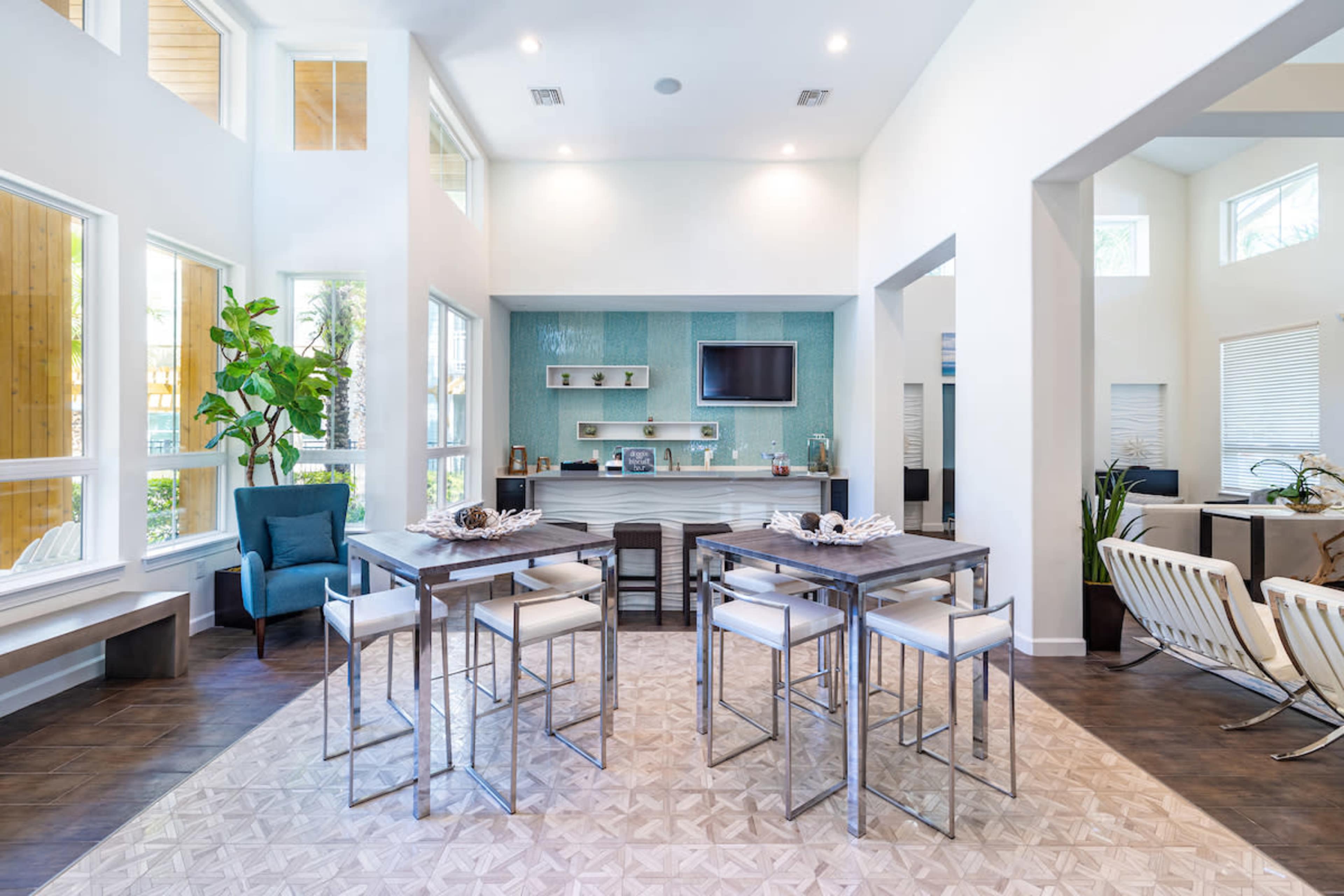 A modern open-concept living space with a dining area featuring tall tables and chairs, and a kitchenette area adorned with light blue tiles and a television.