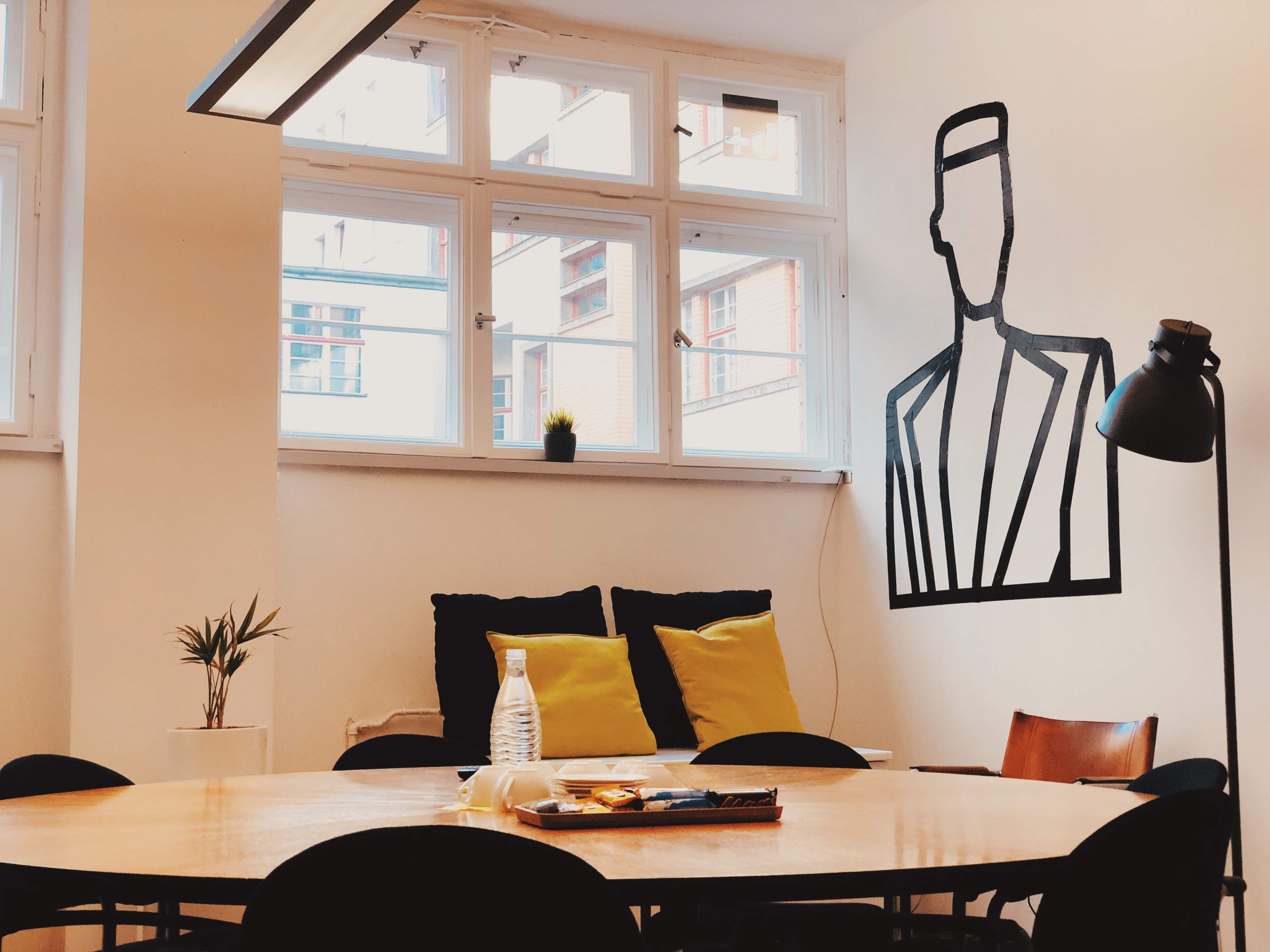 A modern room with a large round table, a black lamp, two yellow cushions on a couch, and a minimalist outline of a man on the wall.