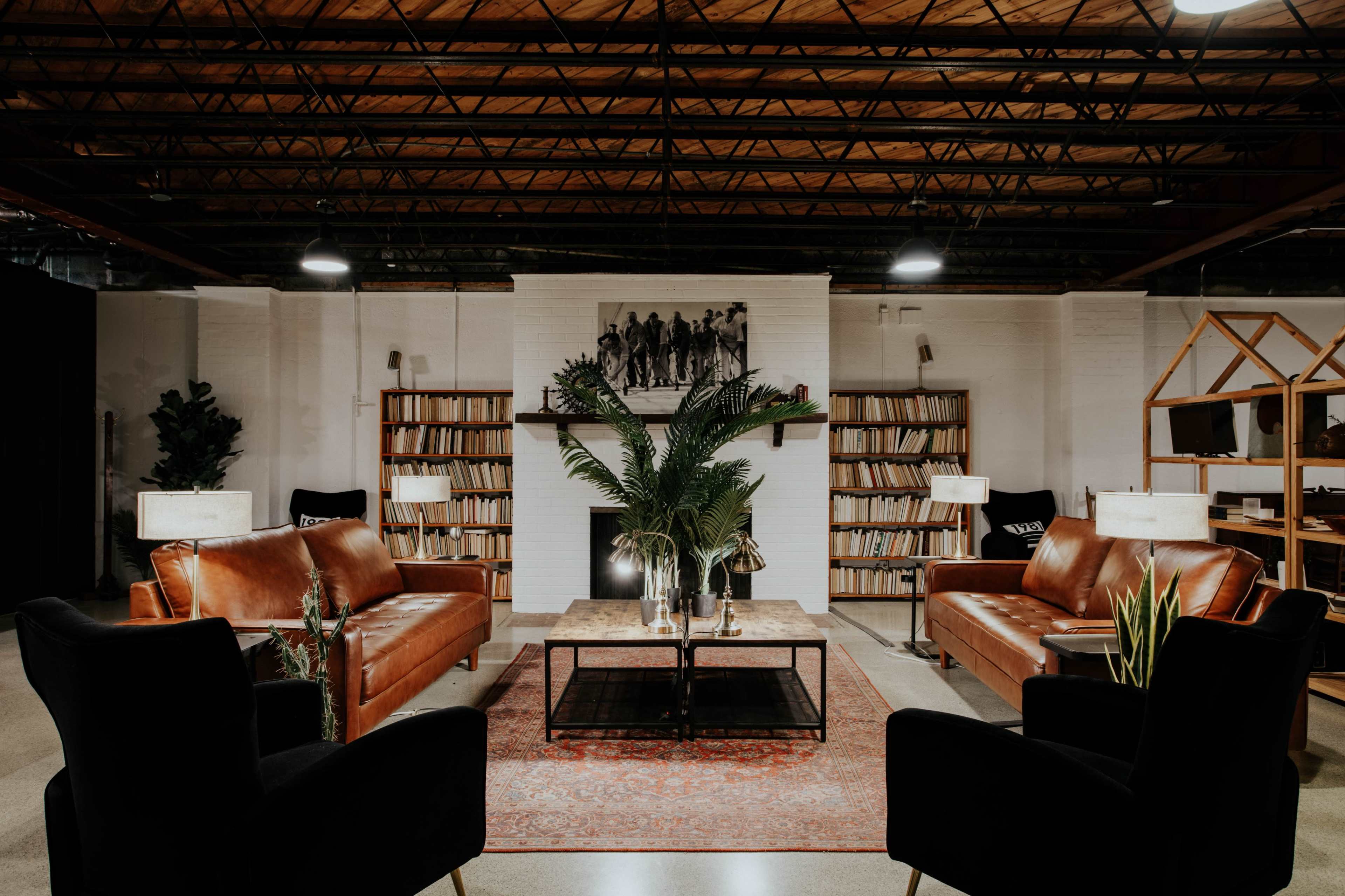 A modern lounge area featuring two brown leather sofas, a glass coffee table, and bookshelves filled with books against a backdrop of a white wall adorned with a decorative arrangement of plants and lamps.