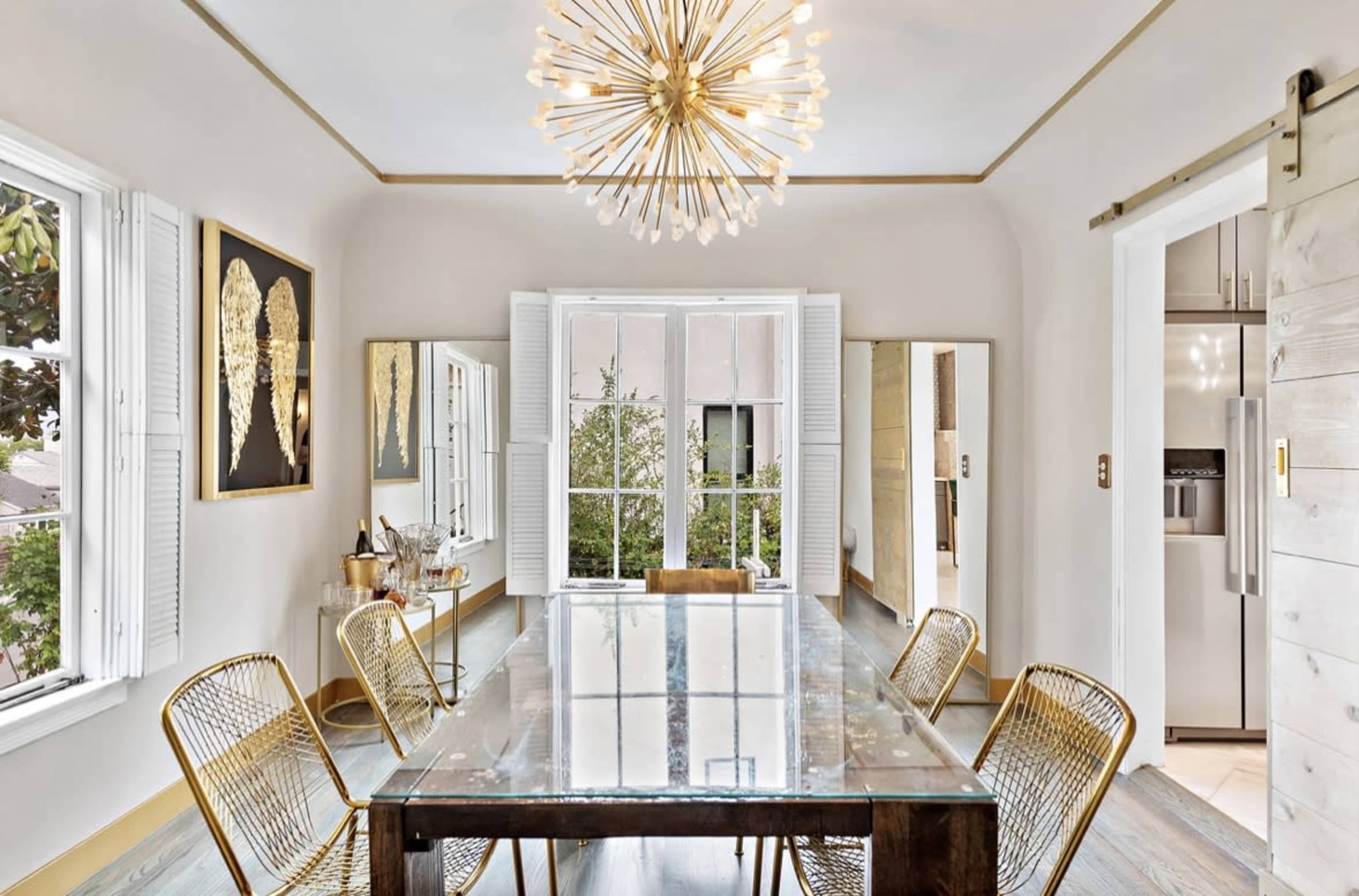 A spacious dining room features a large glass table surrounded by four rattan chairs, with a decorative light fixture overhead and large windows offering a view of greenery outside.