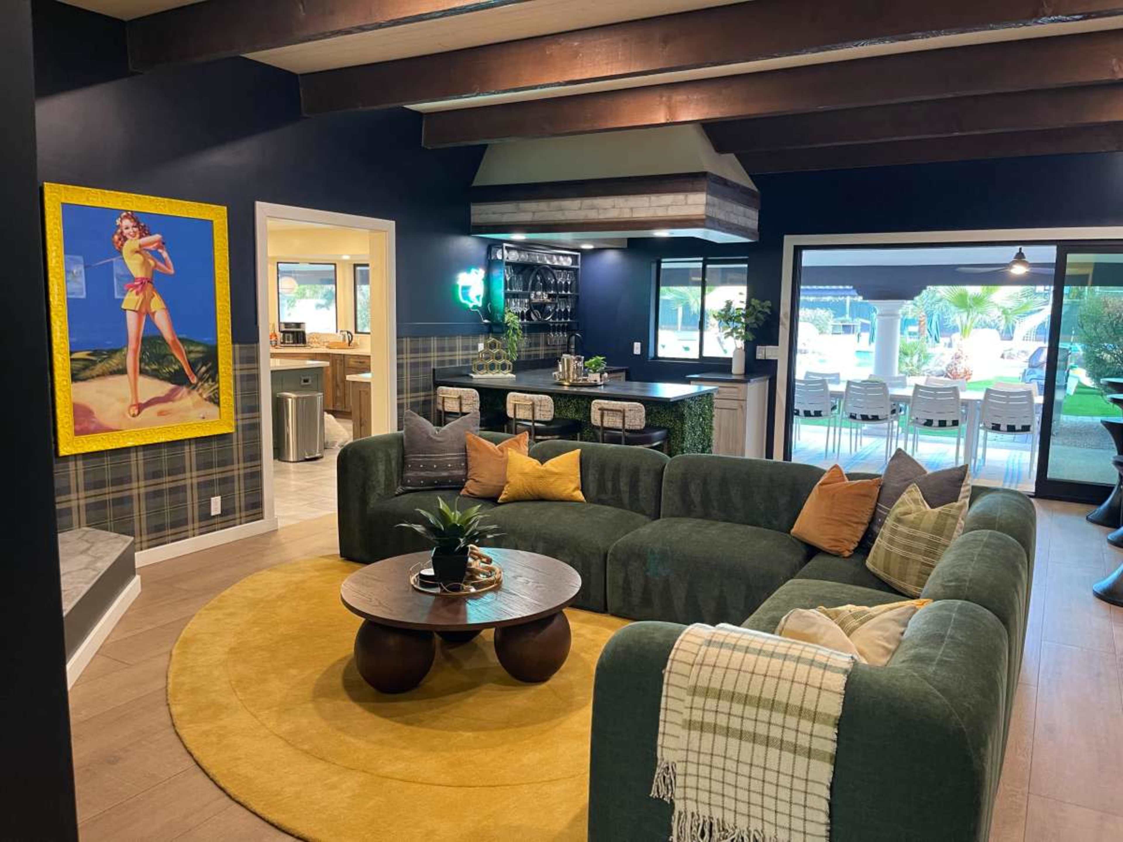 The image showcases a modern living room with a green sectional sofa, a round coffee table, and a large artwork on the wall, with a view of a dining area and outdoor space in the background.