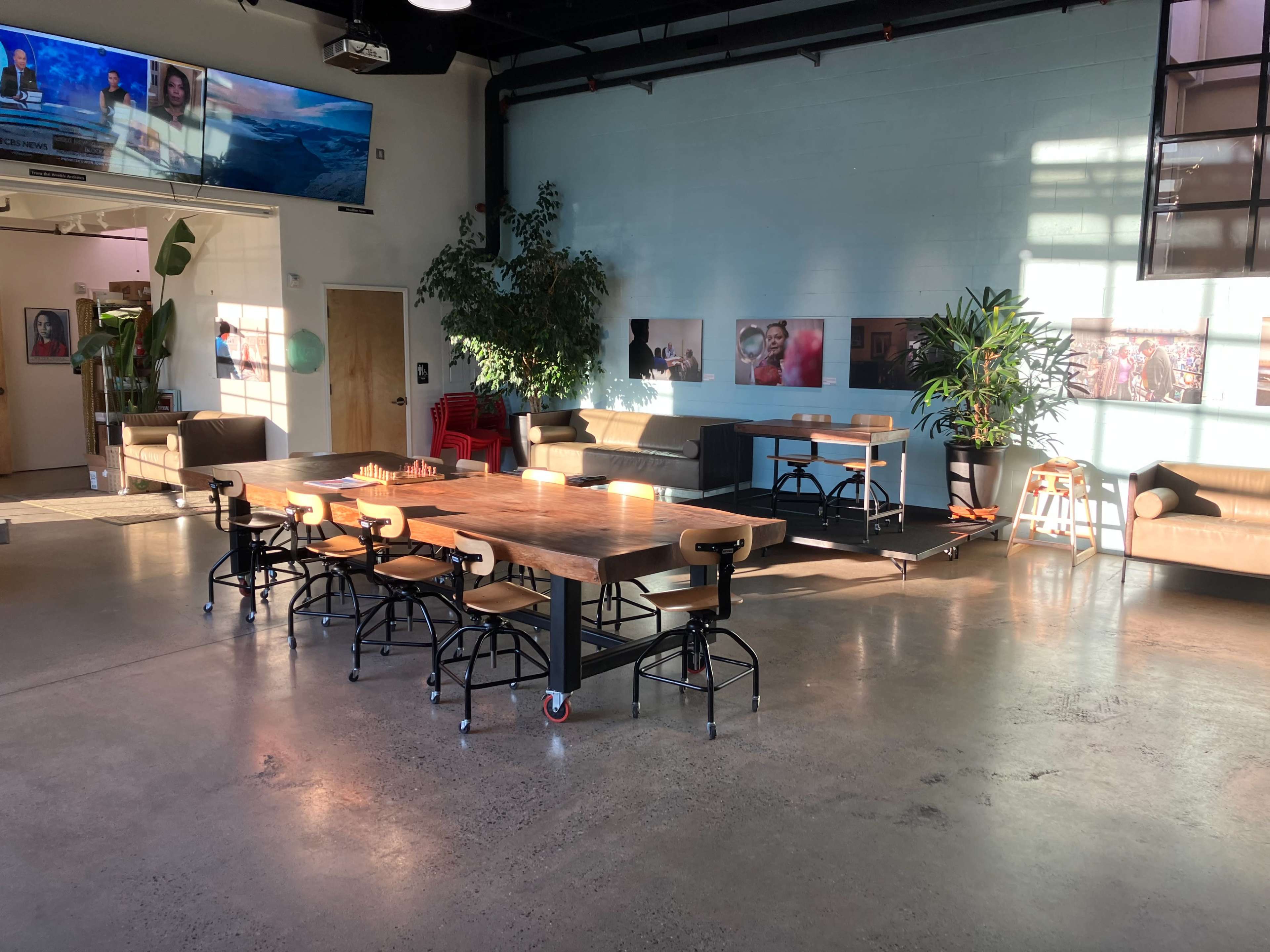 A spacious interior with a large wooden table surrounded by high chairs, plants, and seating areas, illuminated by natural light.