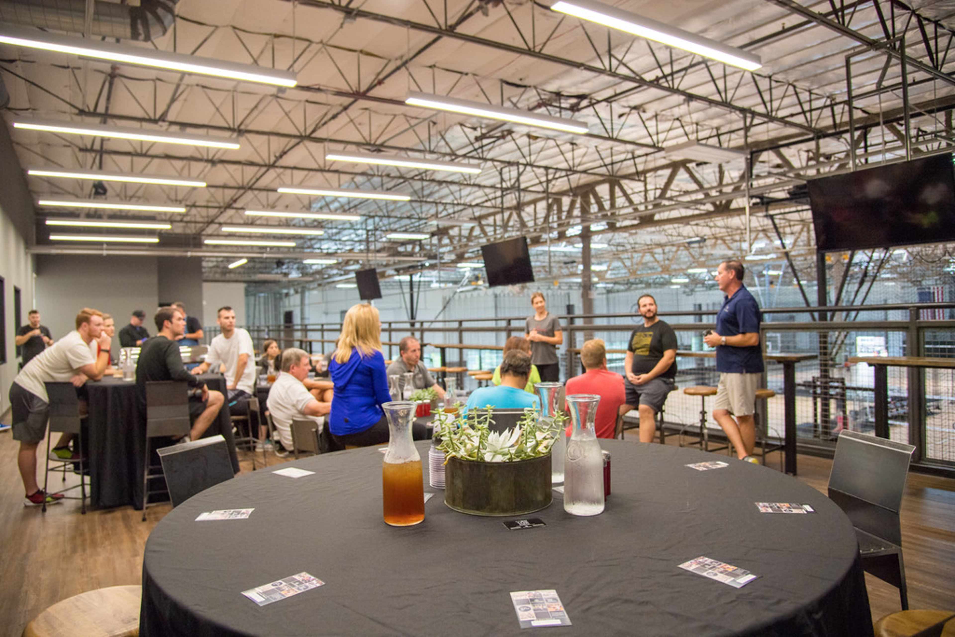 A group of people is seated and standing in a spacious indoor area with high ceilings, engaging in discussion while tables and drinks are arranged around them.