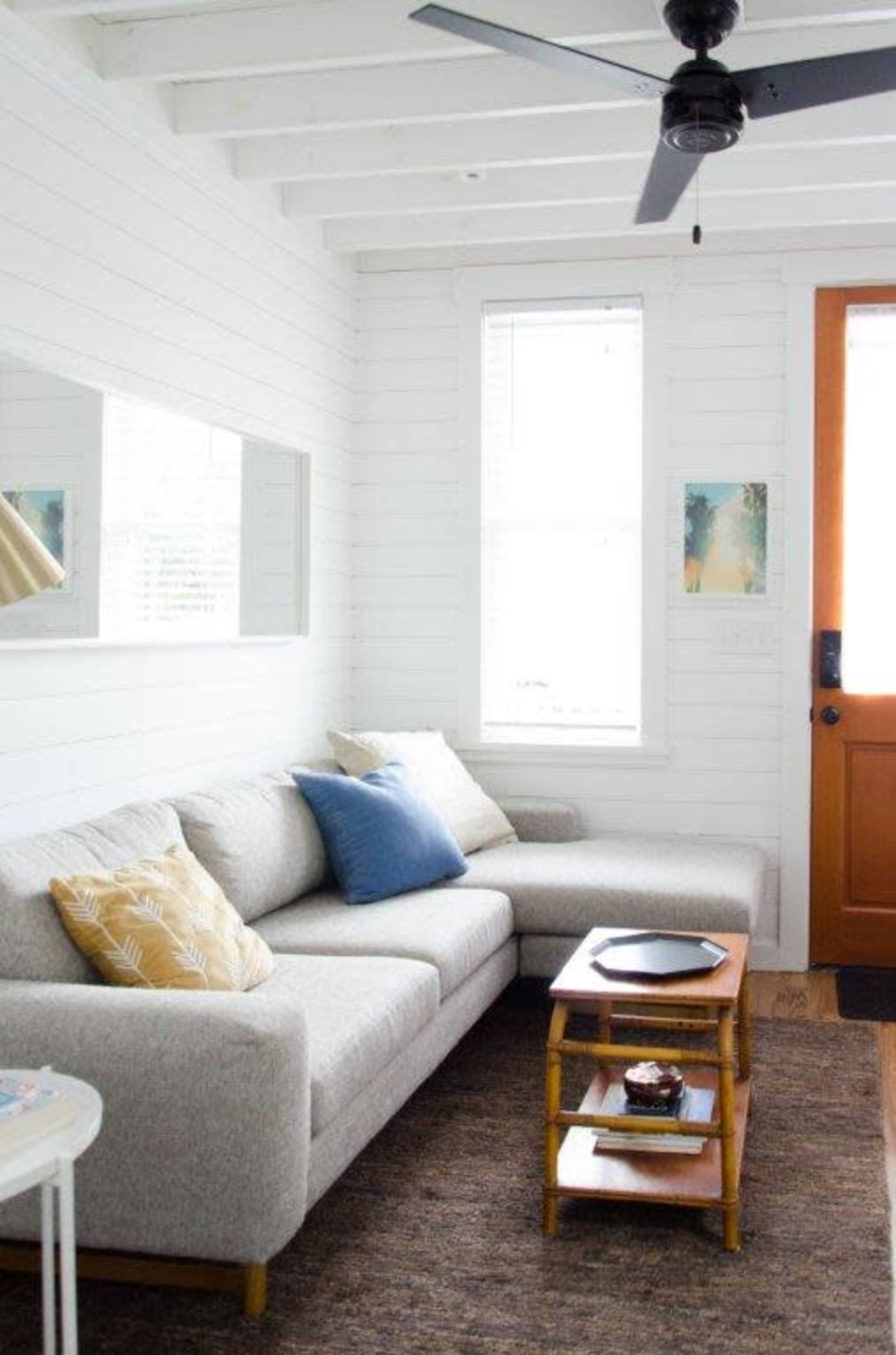 A light-filled living room features a gray sectional sofa with various throw pillows, a wooden coffee table, and a door leading outside.