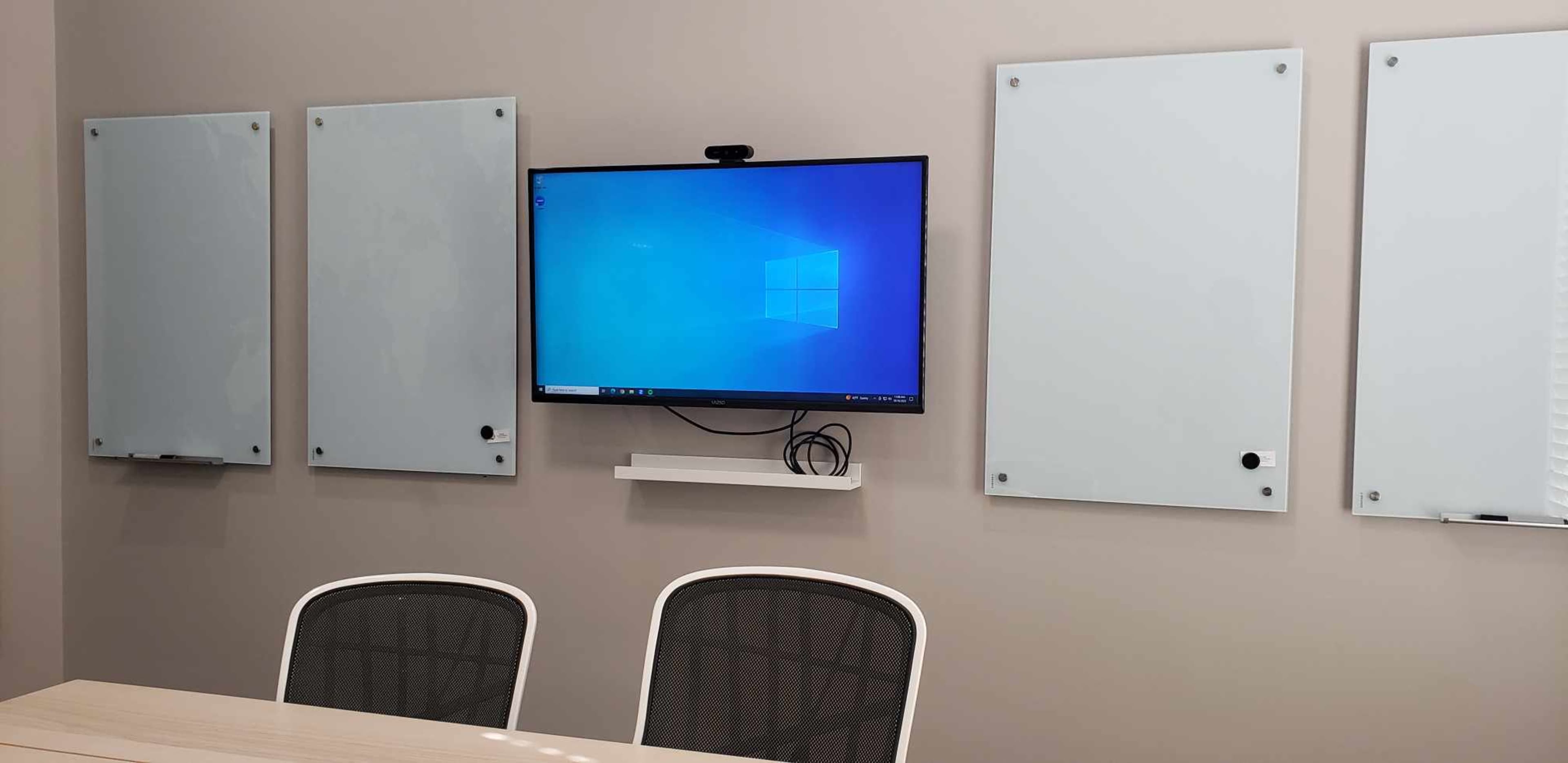 A conference room features a wall-mounted monitor with a Windows desktop, flanked by three blank whiteboards and a table with two chairs.