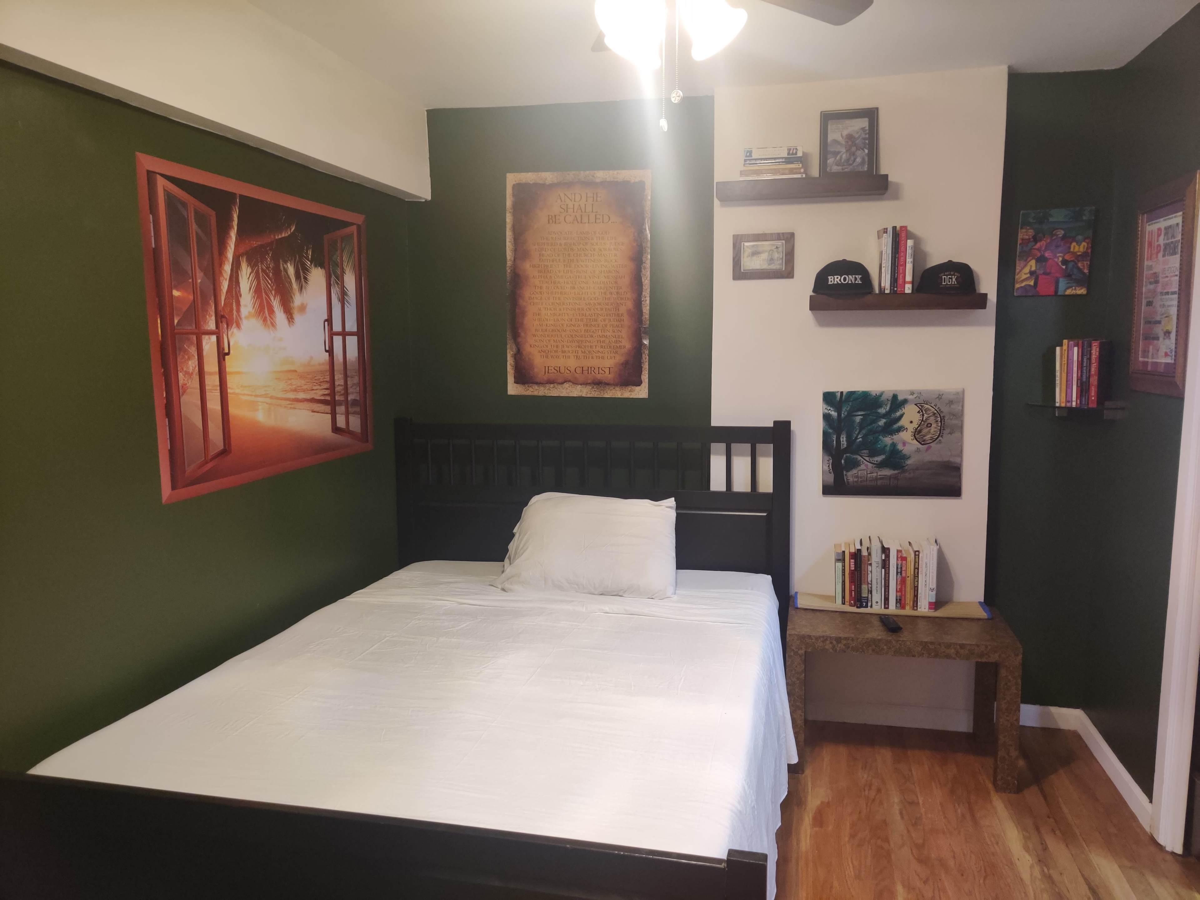 A room with green walls, a black bed with a white bedspread, and several framed artworks and shelves on the walls.