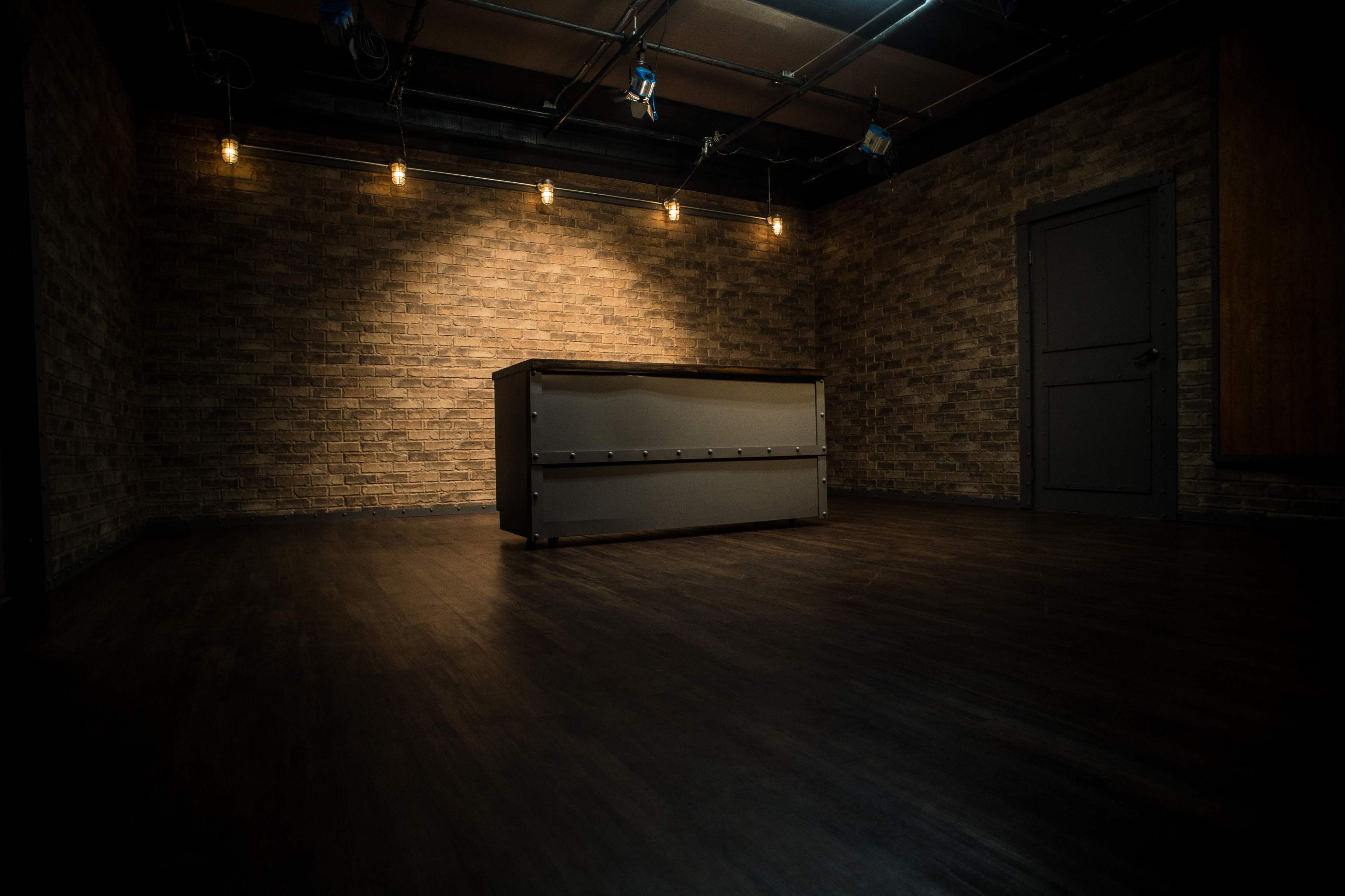 A dimly lit studio space with a stone wall, a dark wooden floor, and a central grey table.