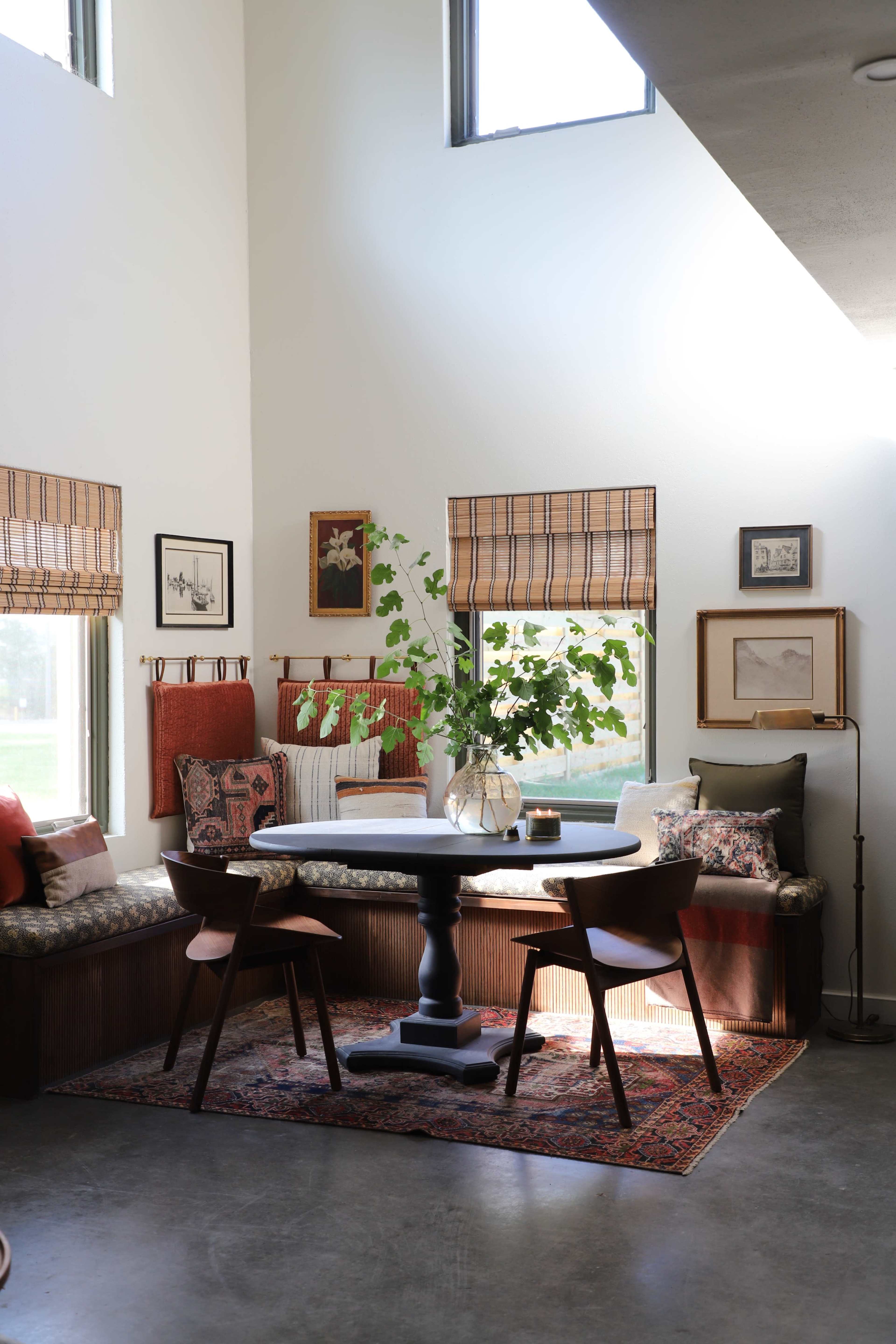 A cozy dining area features a round table with two chairs, surrounded by a window bench adorned with cushions and decorative art on the walls.