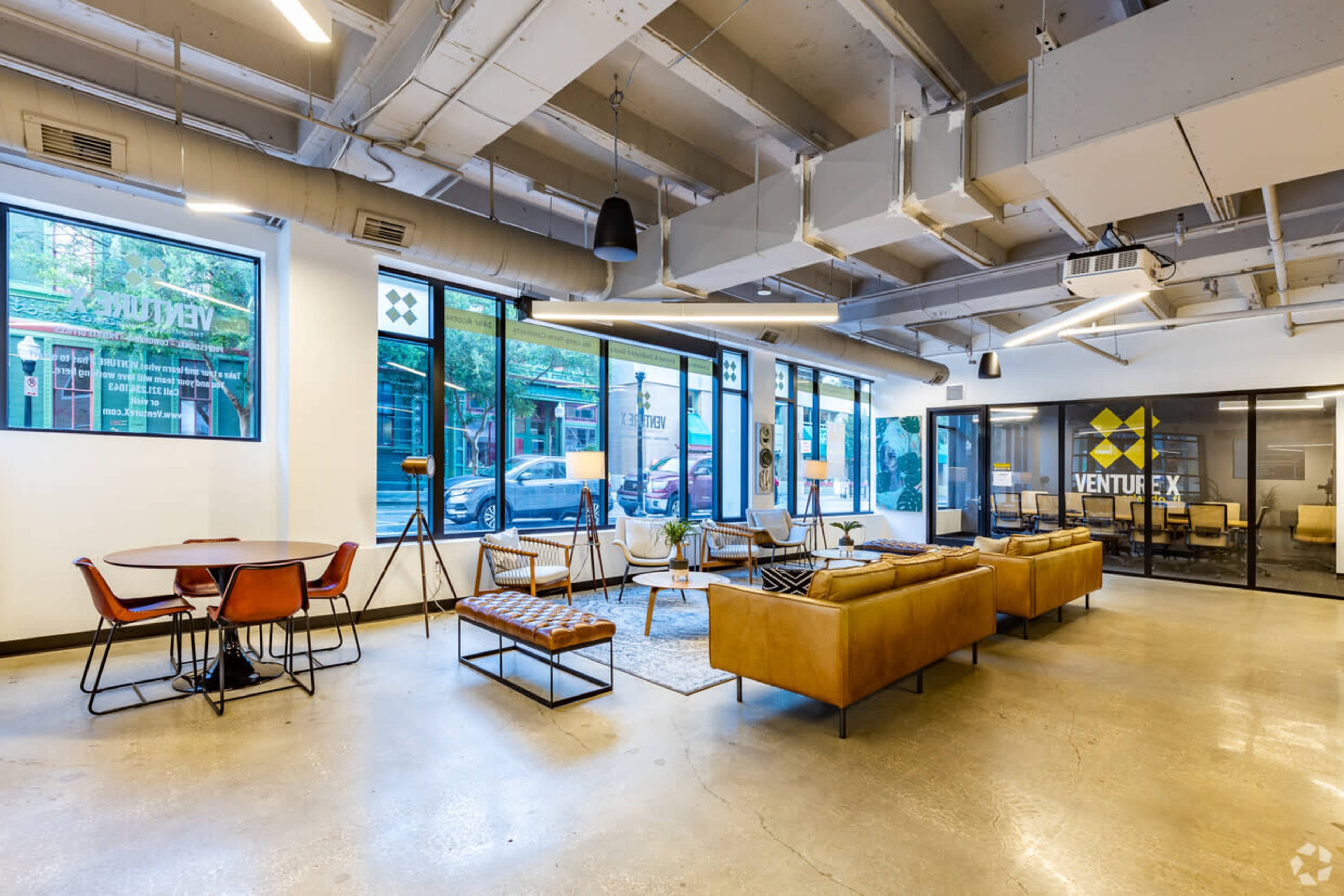 A modern office interior with a seating area featuring brown leather couches, a round dining table, and large windows overlooking the street.