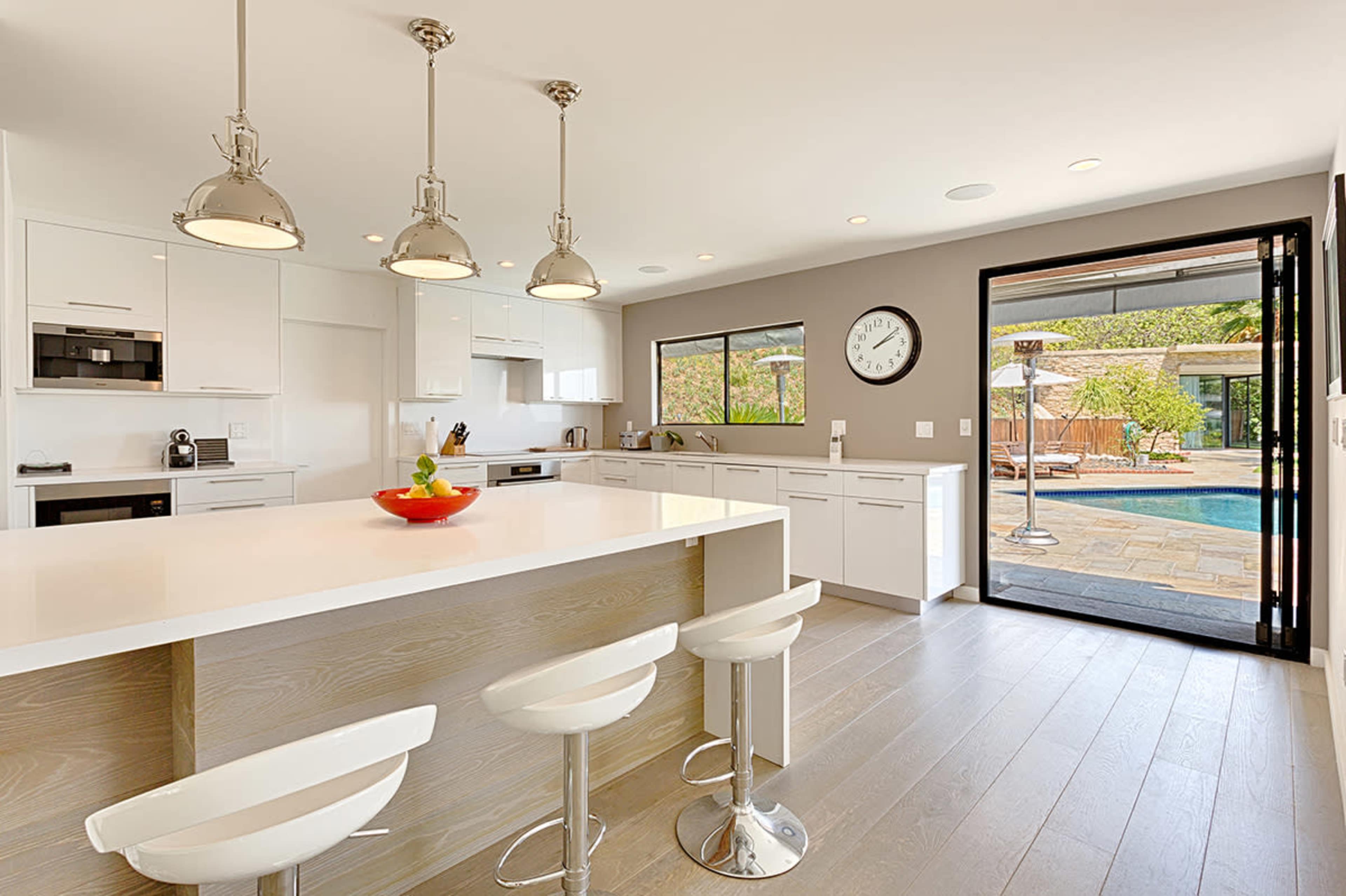 The modern kitchen features white cabinetry, a large island with bar stools, and a sliding door that opens to a pool area.