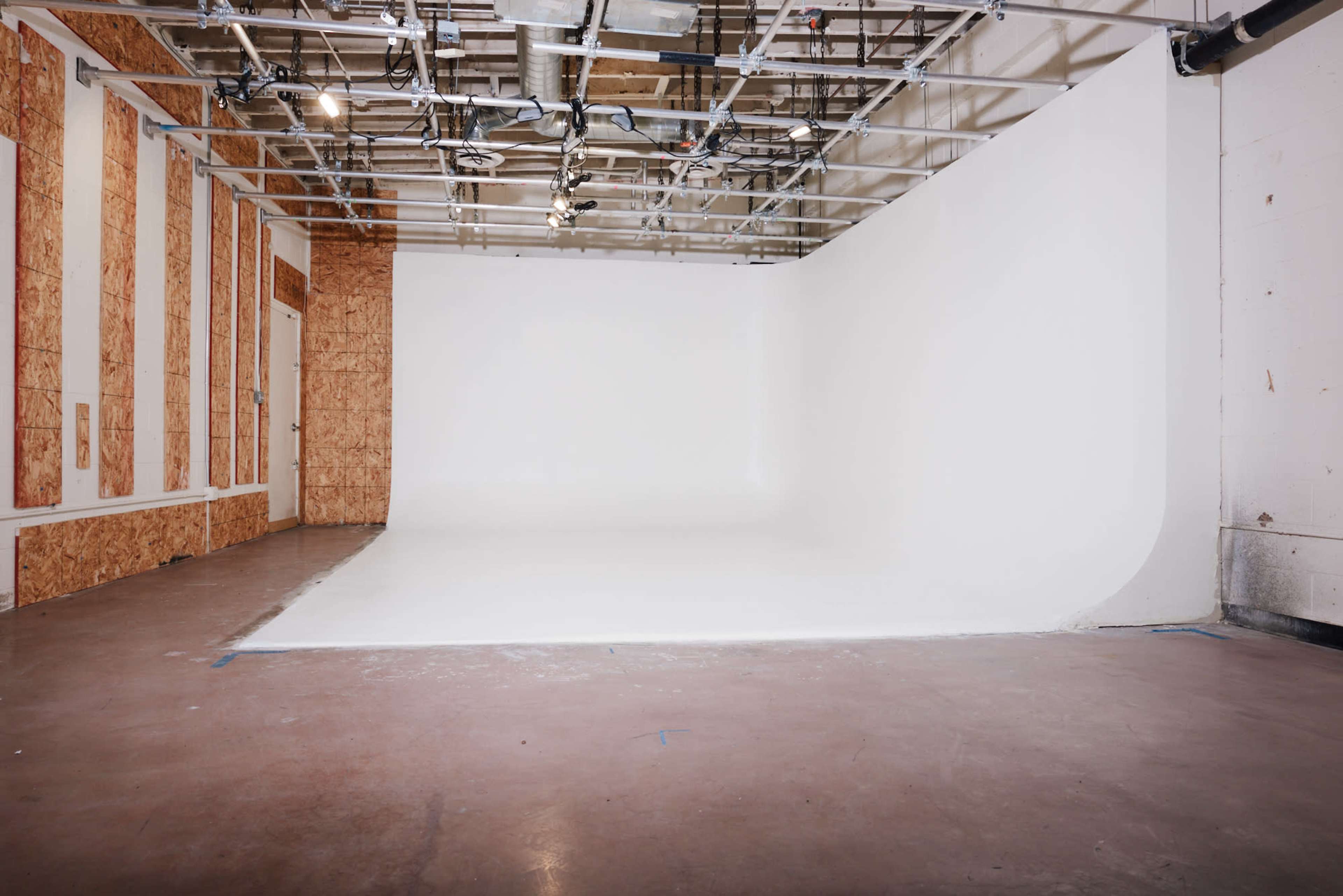 The image shows a spacious, empty studio with a seamless white backdrop, bare walls, and exposed ceiling fixtures.