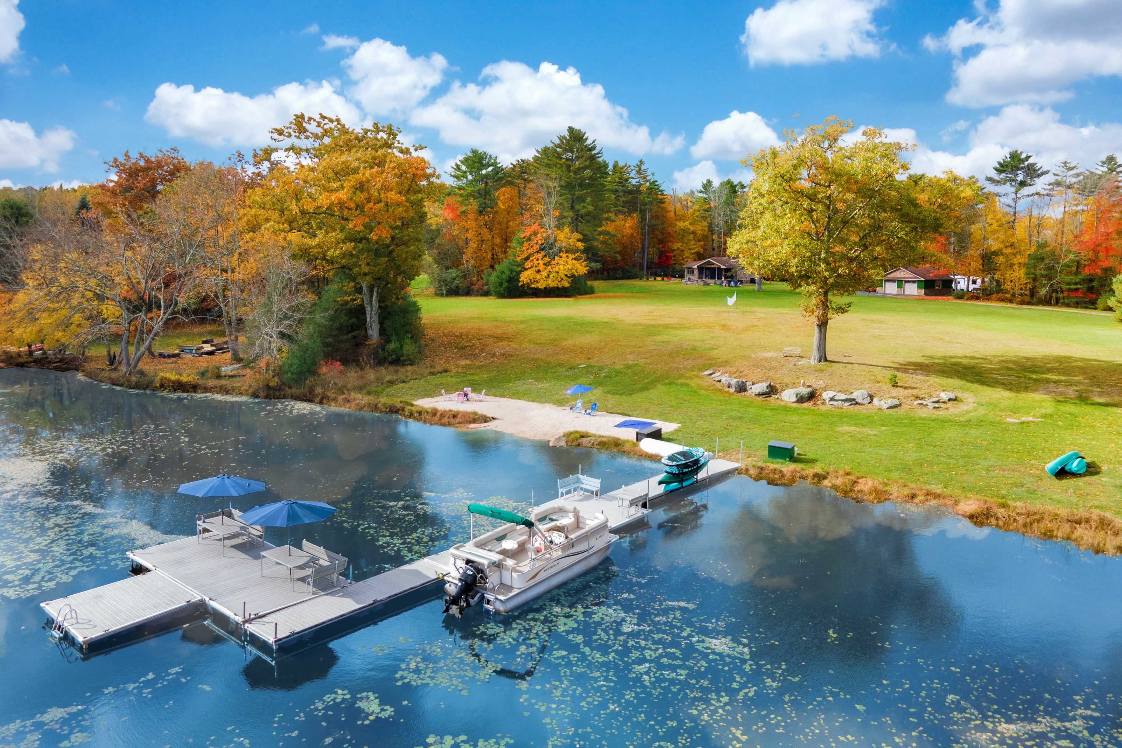 A dock with a boat and umbrellas extends into a calm lake, surrounded by vibrant autumn trees and a grassy area with recreational features.