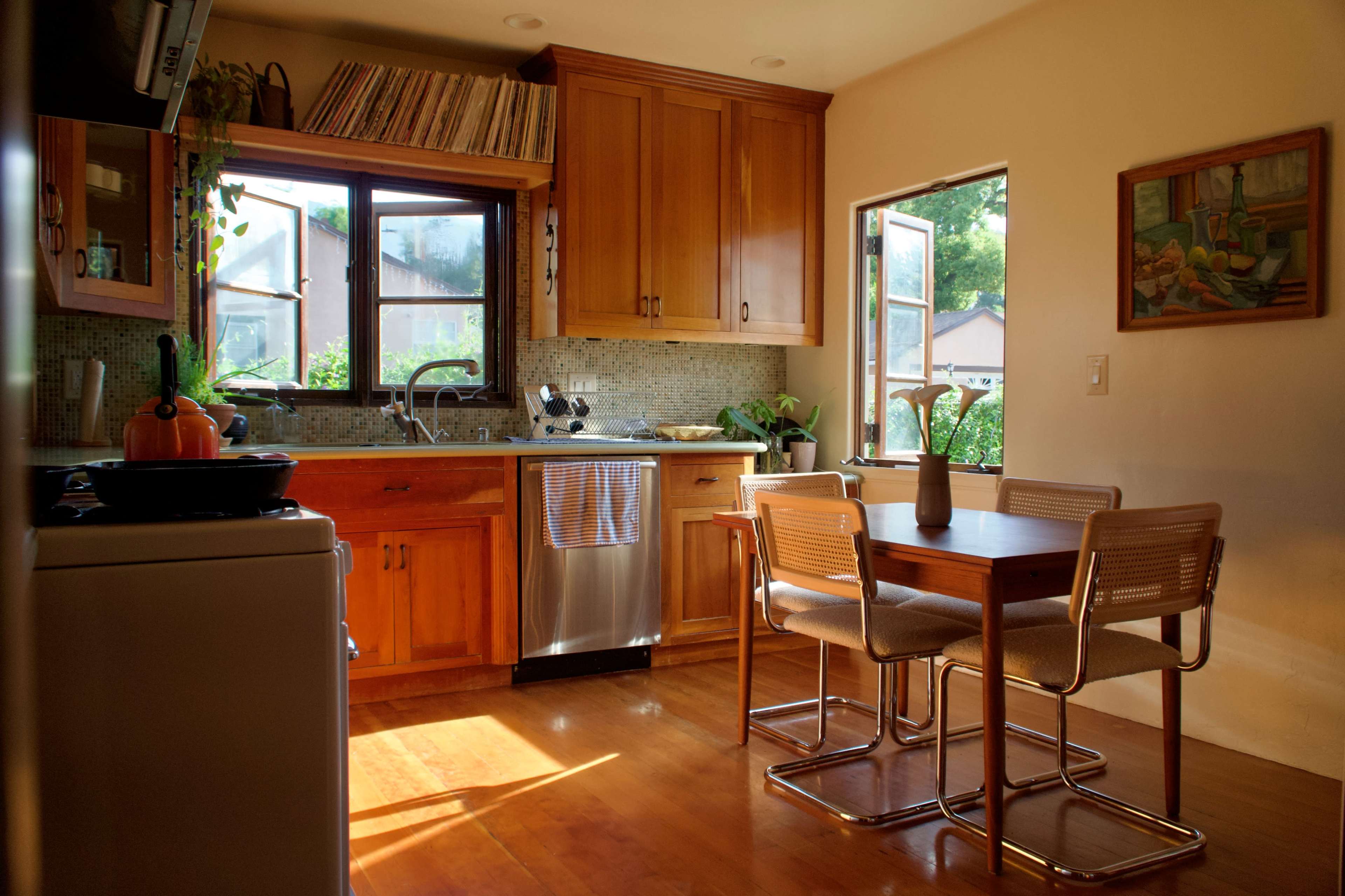 A warm, sunlit kitchen features wooden cabinets, a dinette set with four chairs, and a window that overlooks a garden.