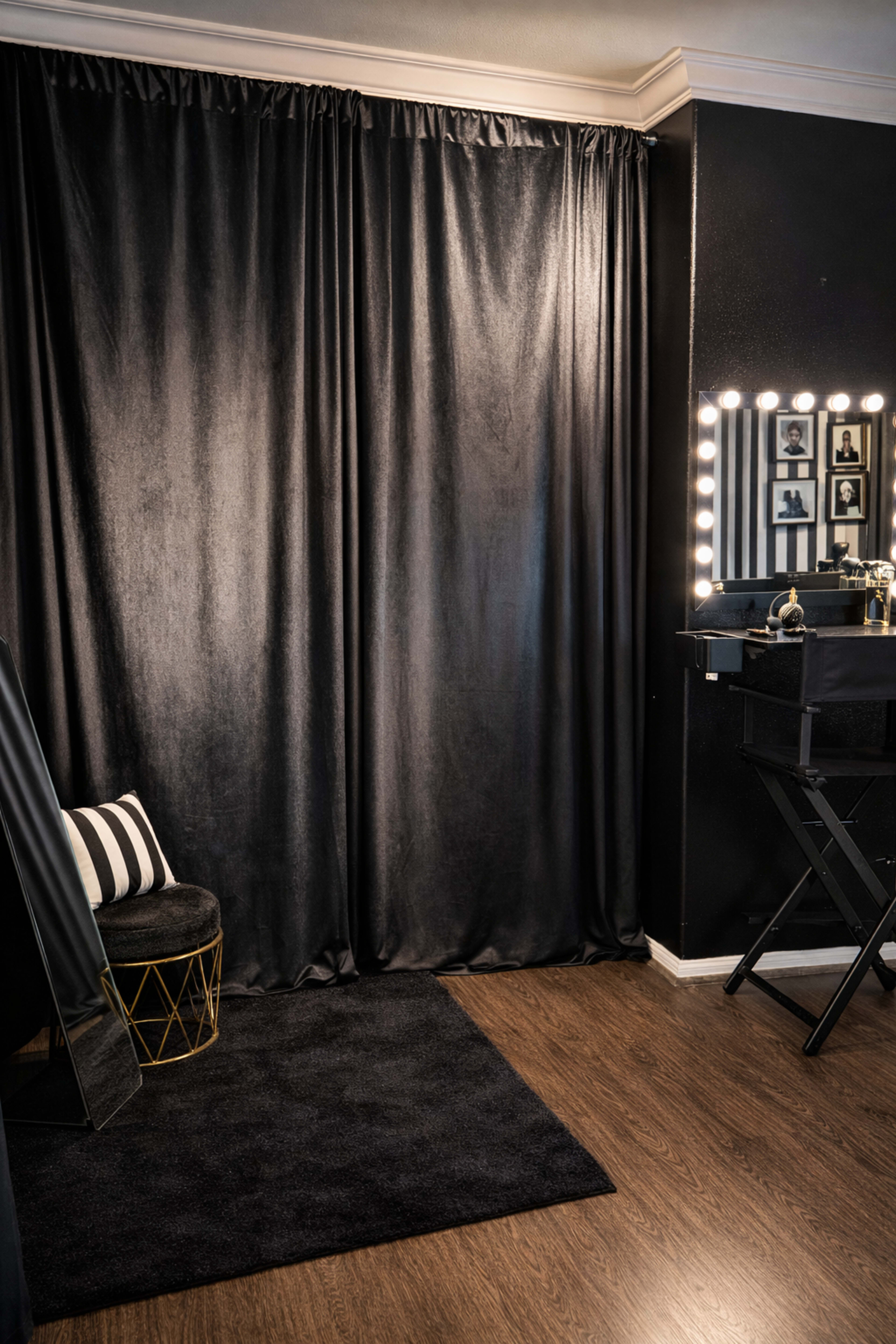 The image shows a dimly lit room featuring a black curtain, a mirror with lighted bulbs, a dark vanity table, and a plush black rug on a wooden floor.