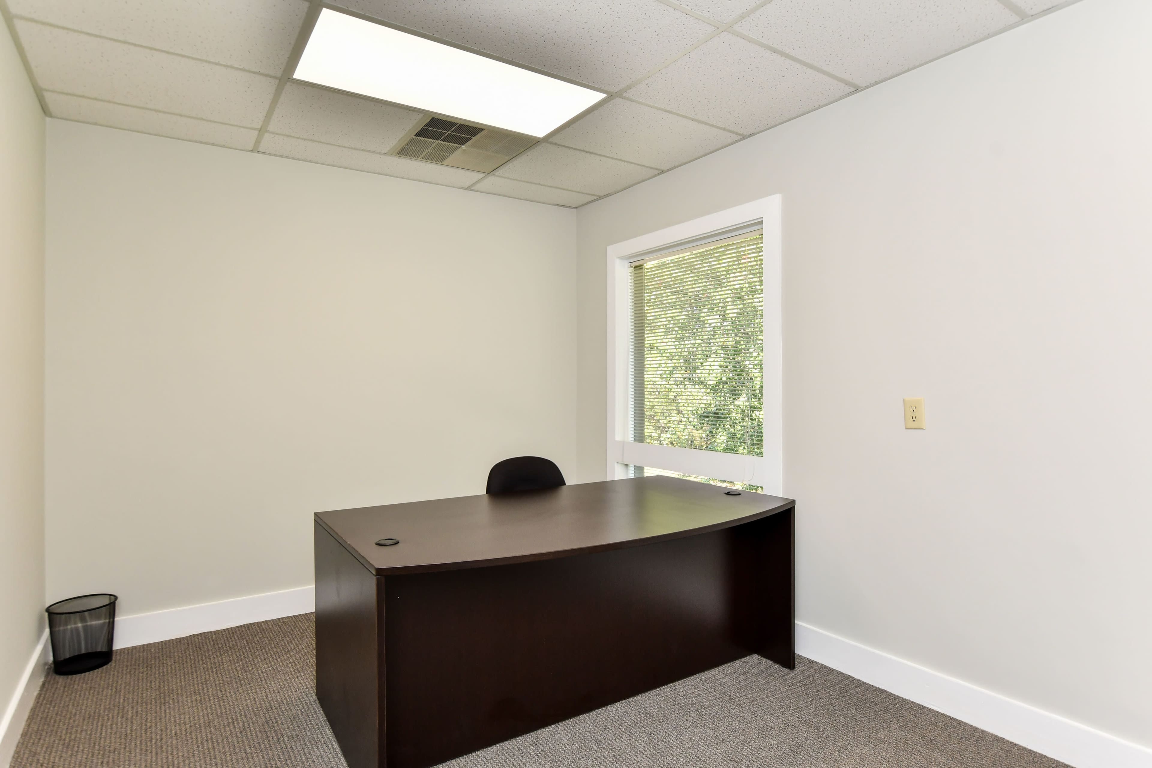 A minimalistic office space featuring a dark wooden desk, a chair, a wastebasket, and a window with blinds.