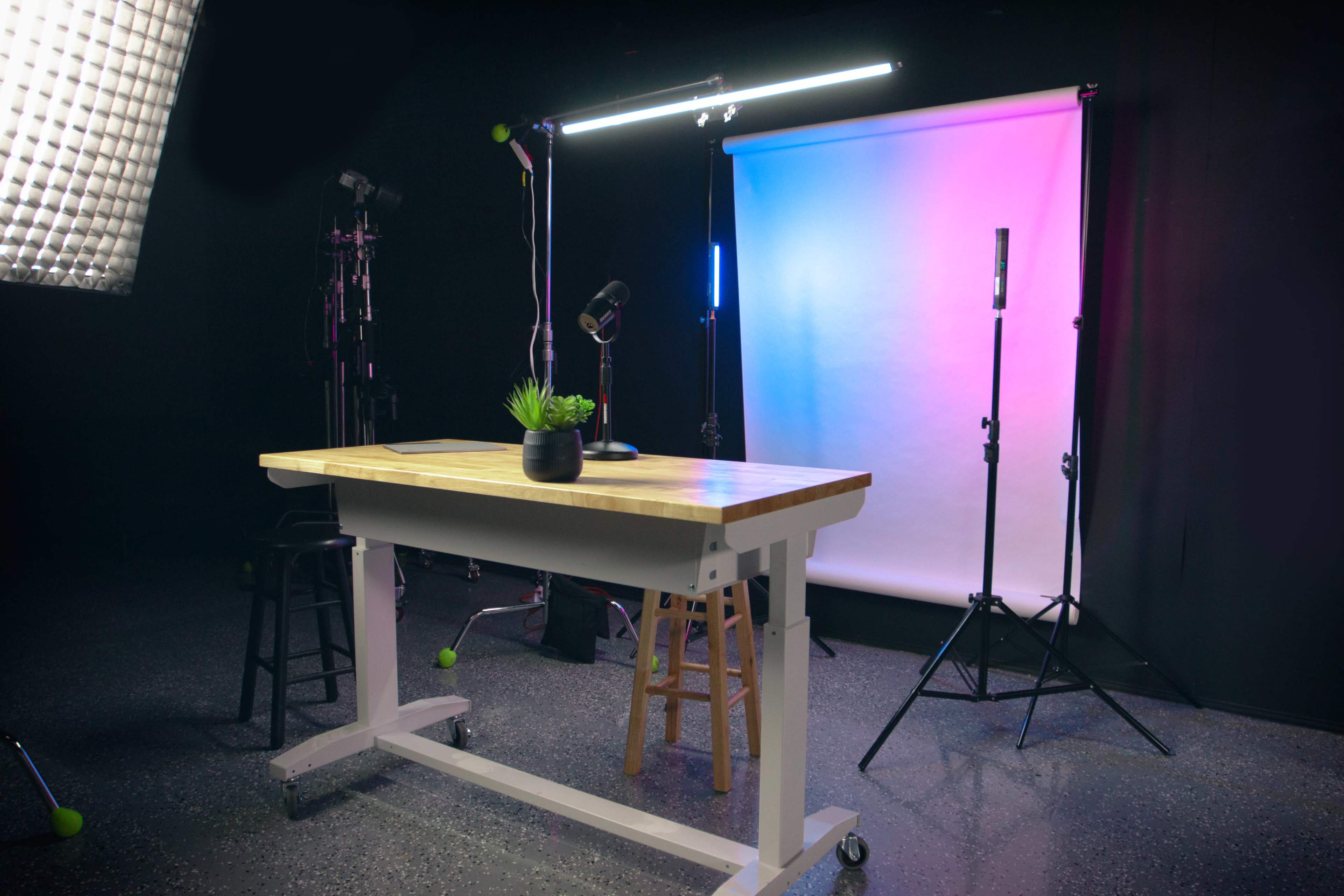 The image shows a well-equipped studio setup featuring a table, a potted plant, lighting equipment, and a backdrop with gradient colors.