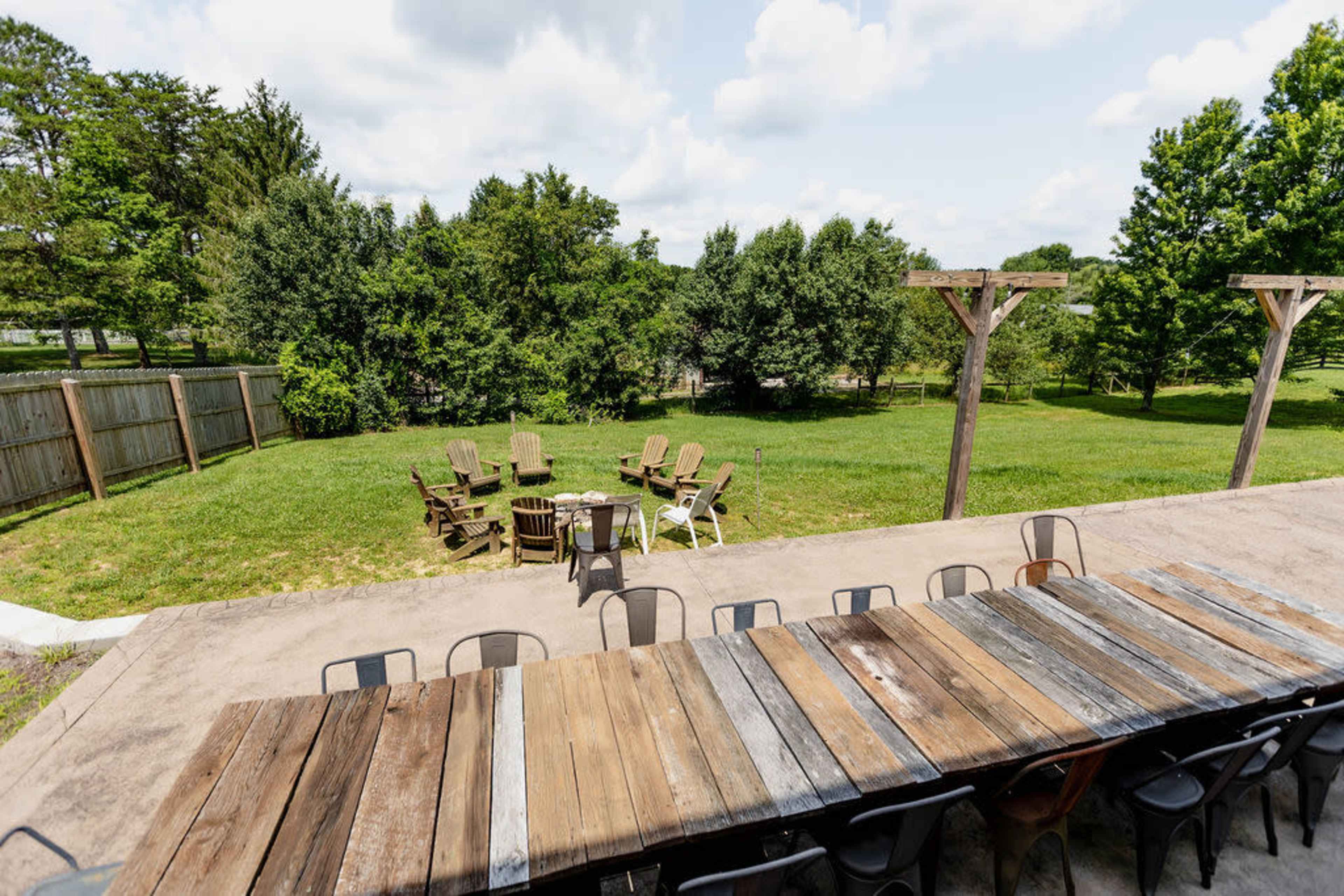 The image shows an outdoor gathering space with a long wooden table and several Adirondack chairs arranged in a circular formation on a grassy area, surrounded by trees.