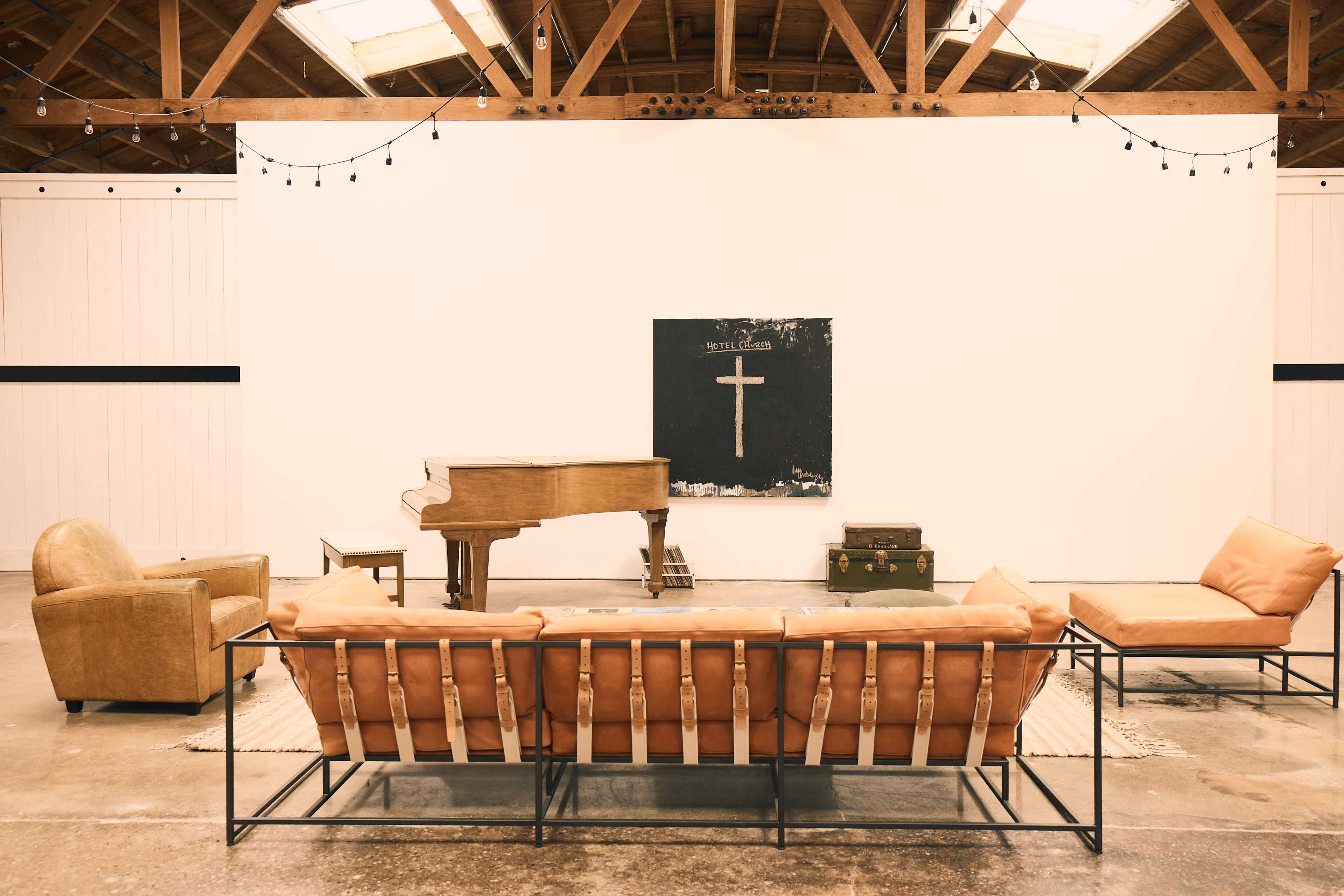The image shows a minimalist interior space featuring a sofa, armchair, a grand piano, and a large blackboard with a cross painted on it.