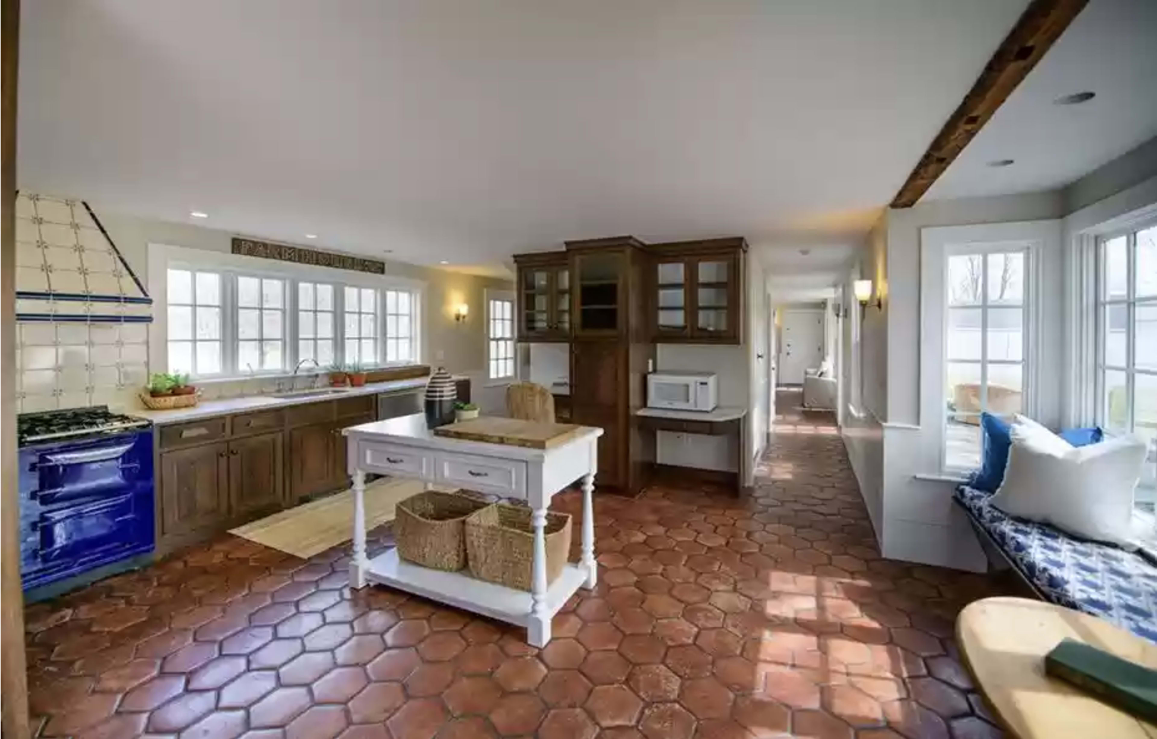 The image shows a spacious kitchen with a blue stove, wooden cabinets, a central island, and large windows that provide ample natural light.