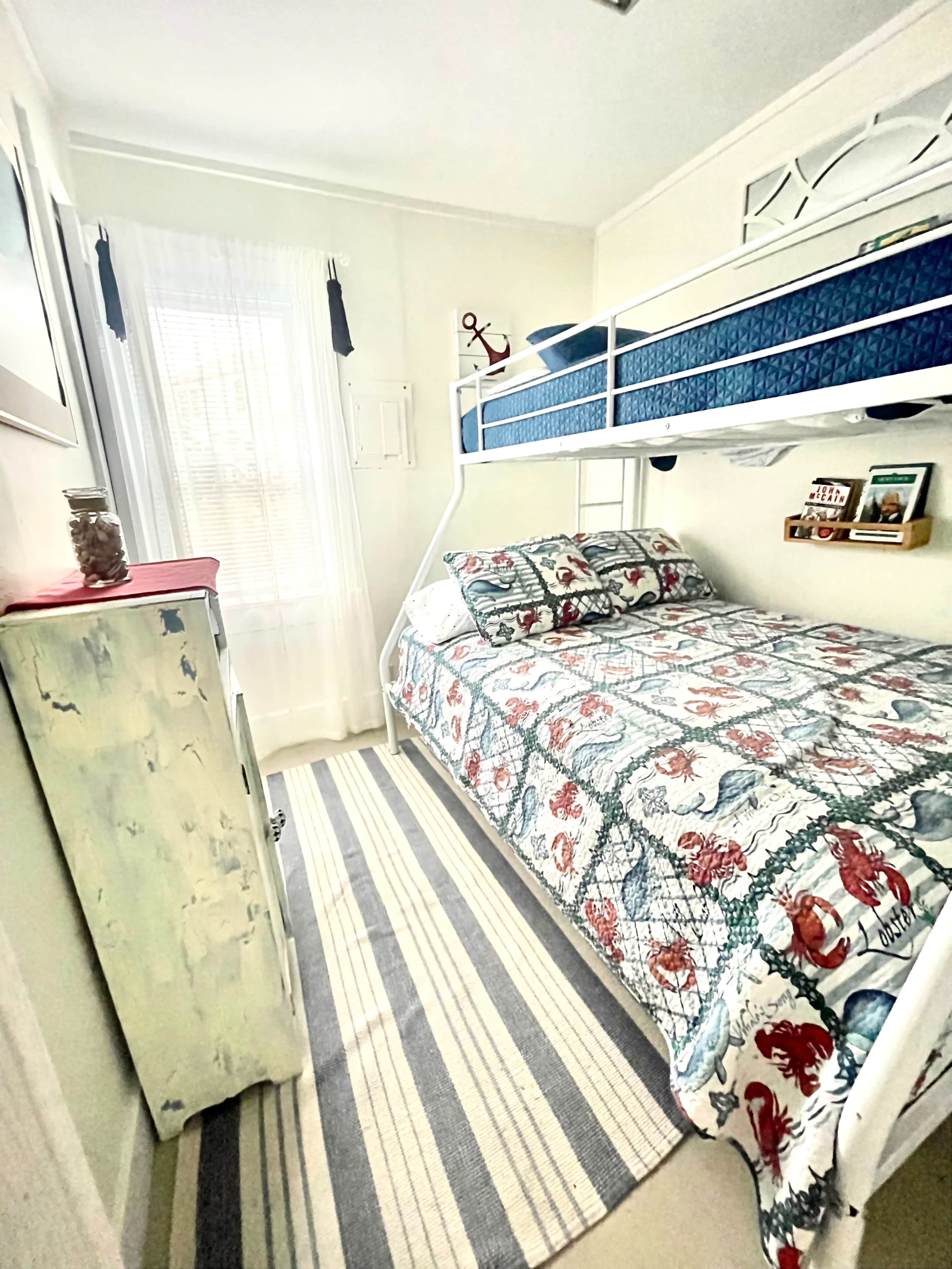 A small bedroom featuring a bunk bed with blue bedding, a lower double bed with a patterned blanket, and a striped area rug on the floor.