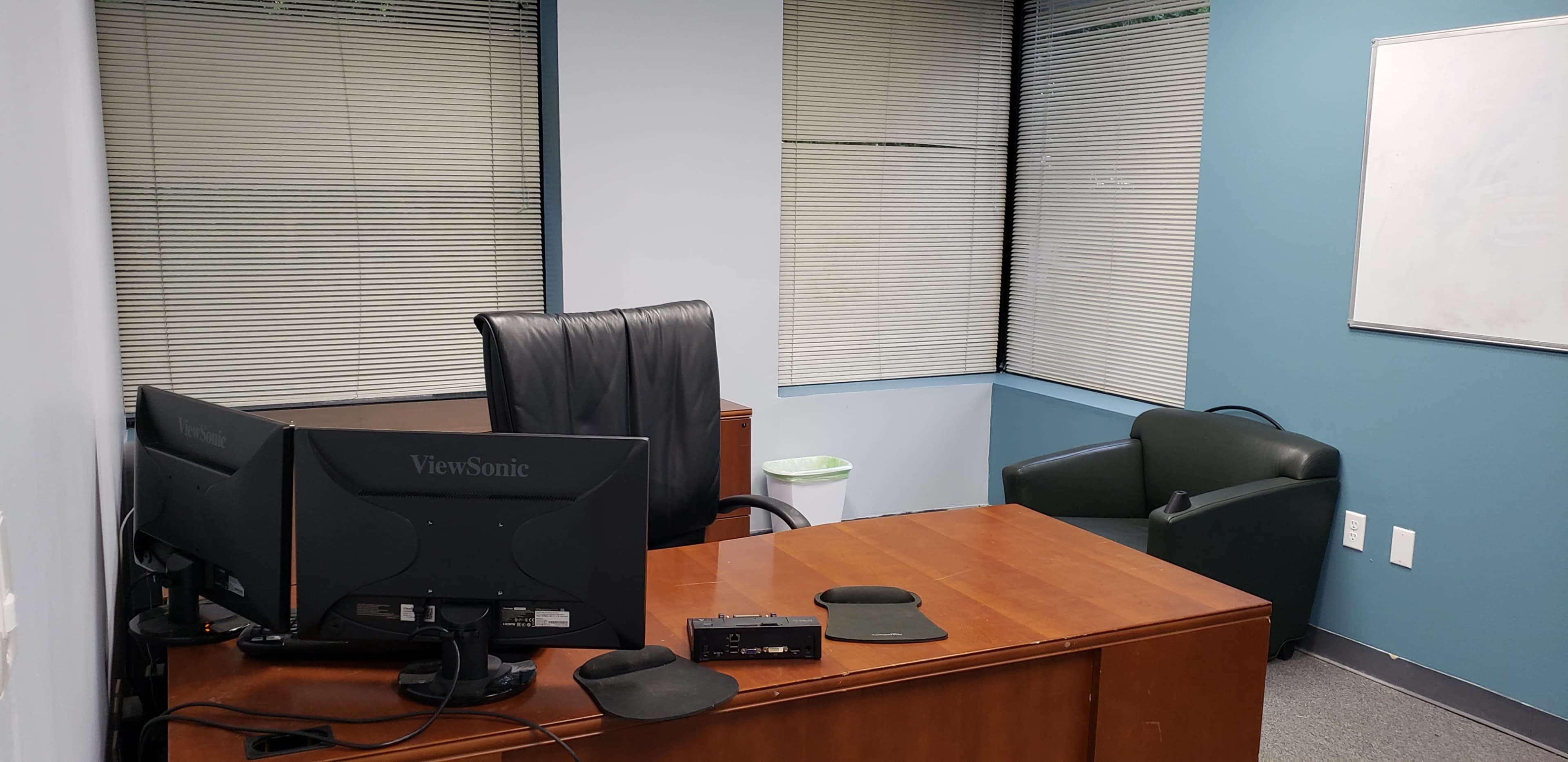 A small office with a wooden desk, an office chair, a computer monitor, and a green armchair beside two windows with blinds.
