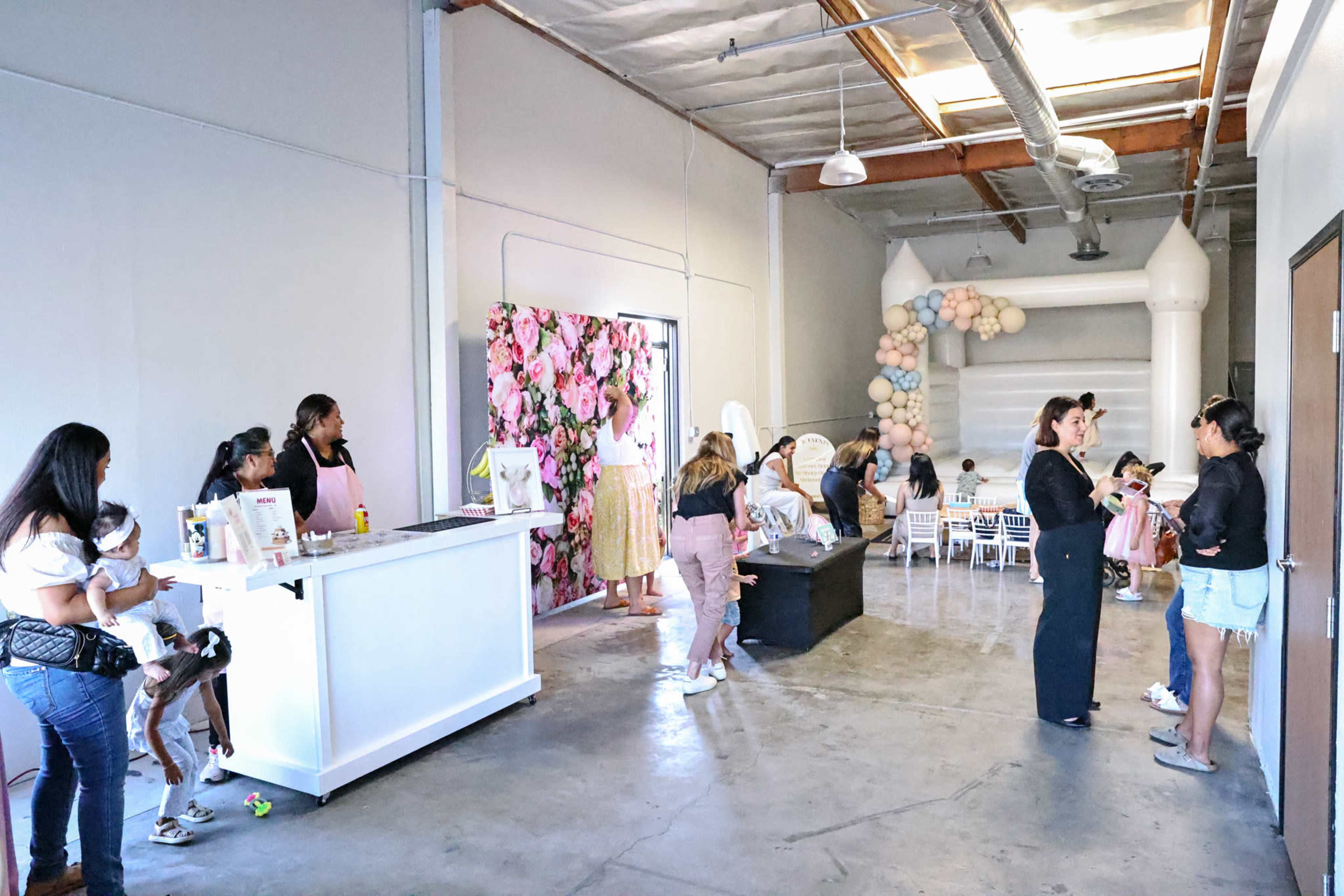 A group of people are gathered inside an event space featuring a floral wall, a balloon arch, and a bouncy castle in the background.