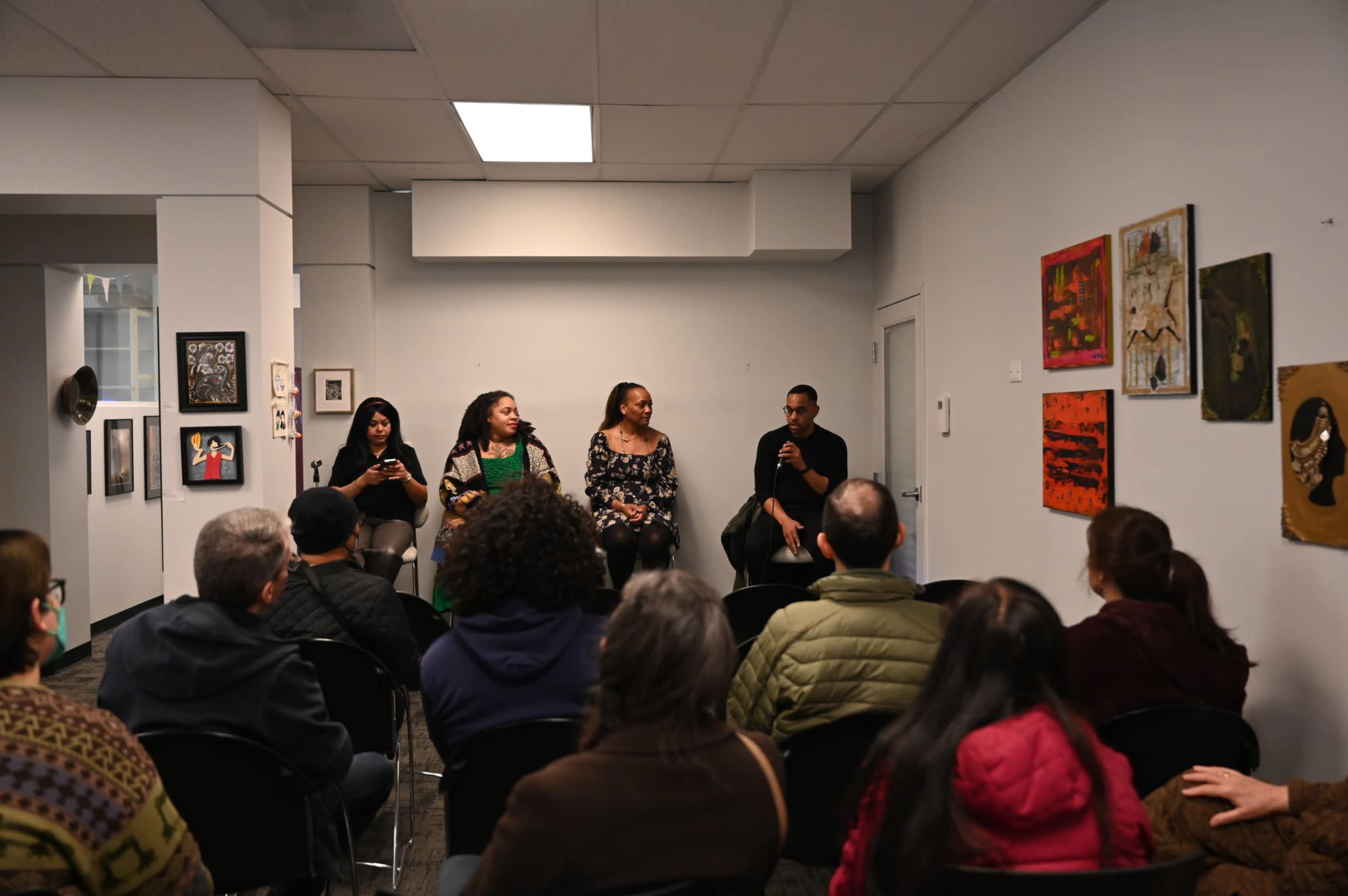 A panel discussion is taking place in a modern room, featuring four speakers seated at the front while an audience listens attentively.