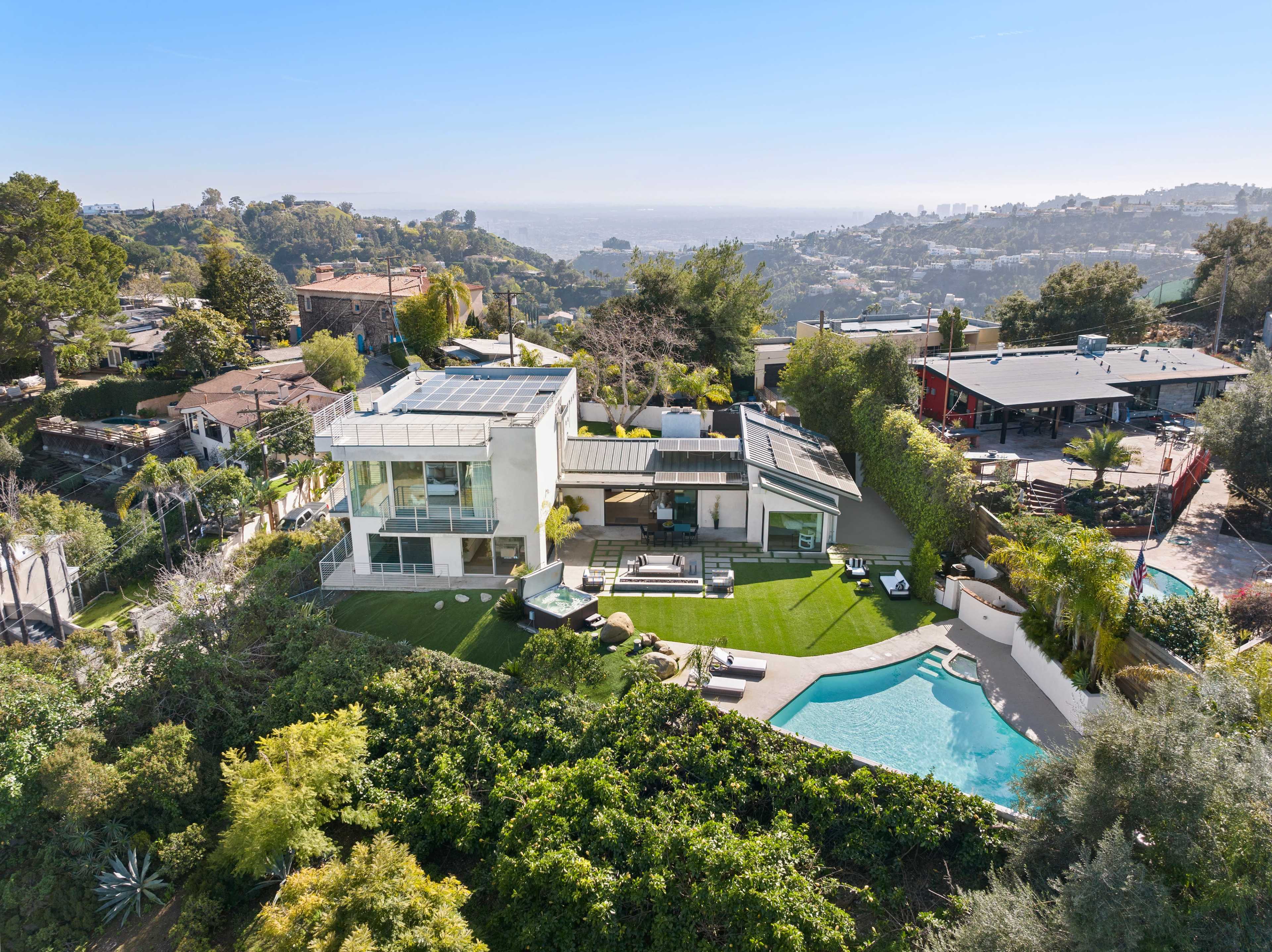 A modern house with a pool is situated on a hillside, surrounded by greenery and offering a view of the landscape and distant hills.