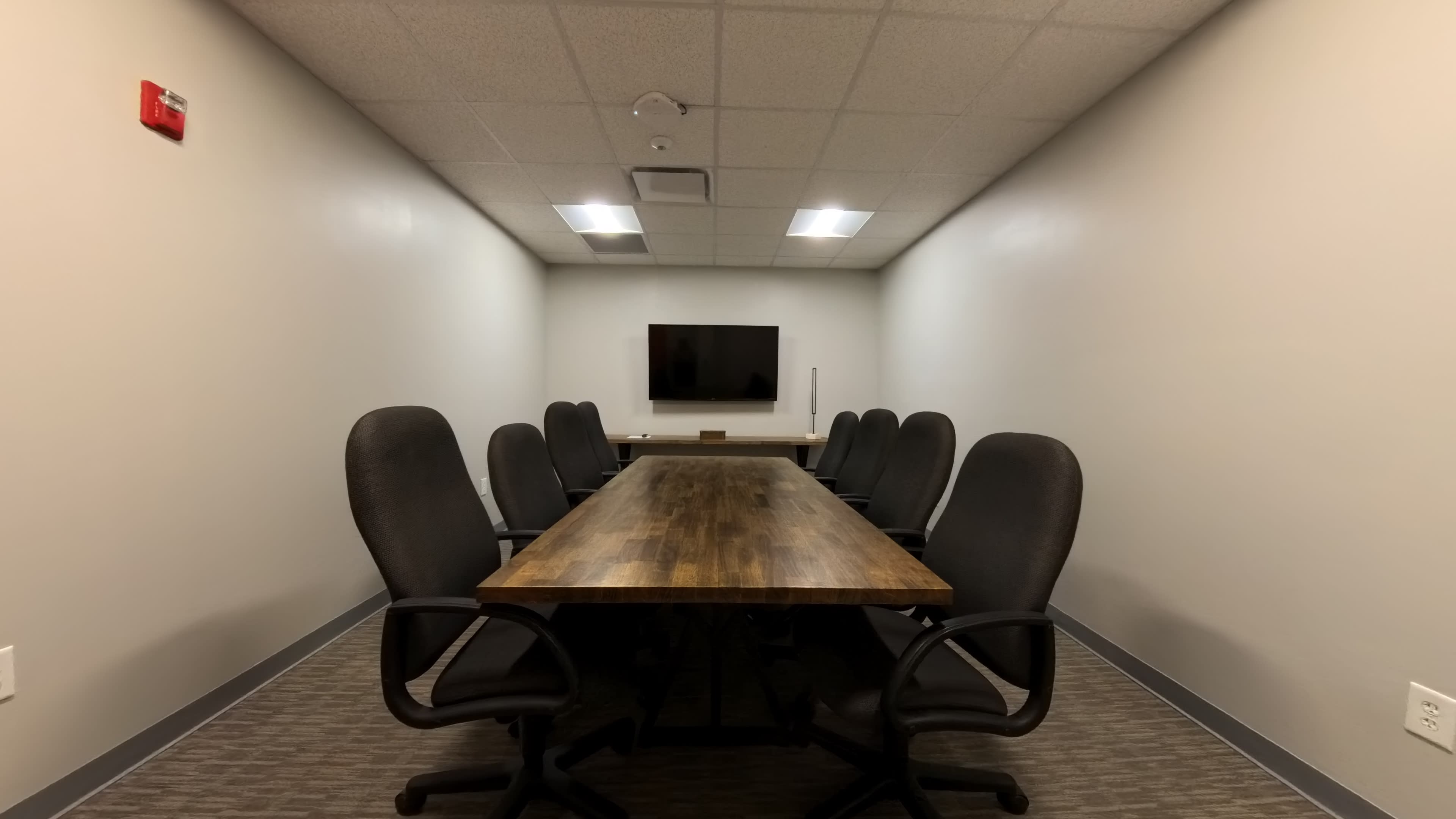 A long wooden conference table with eight black office chairs is set in a small meeting room featuring a flat-screen TV on the wall.