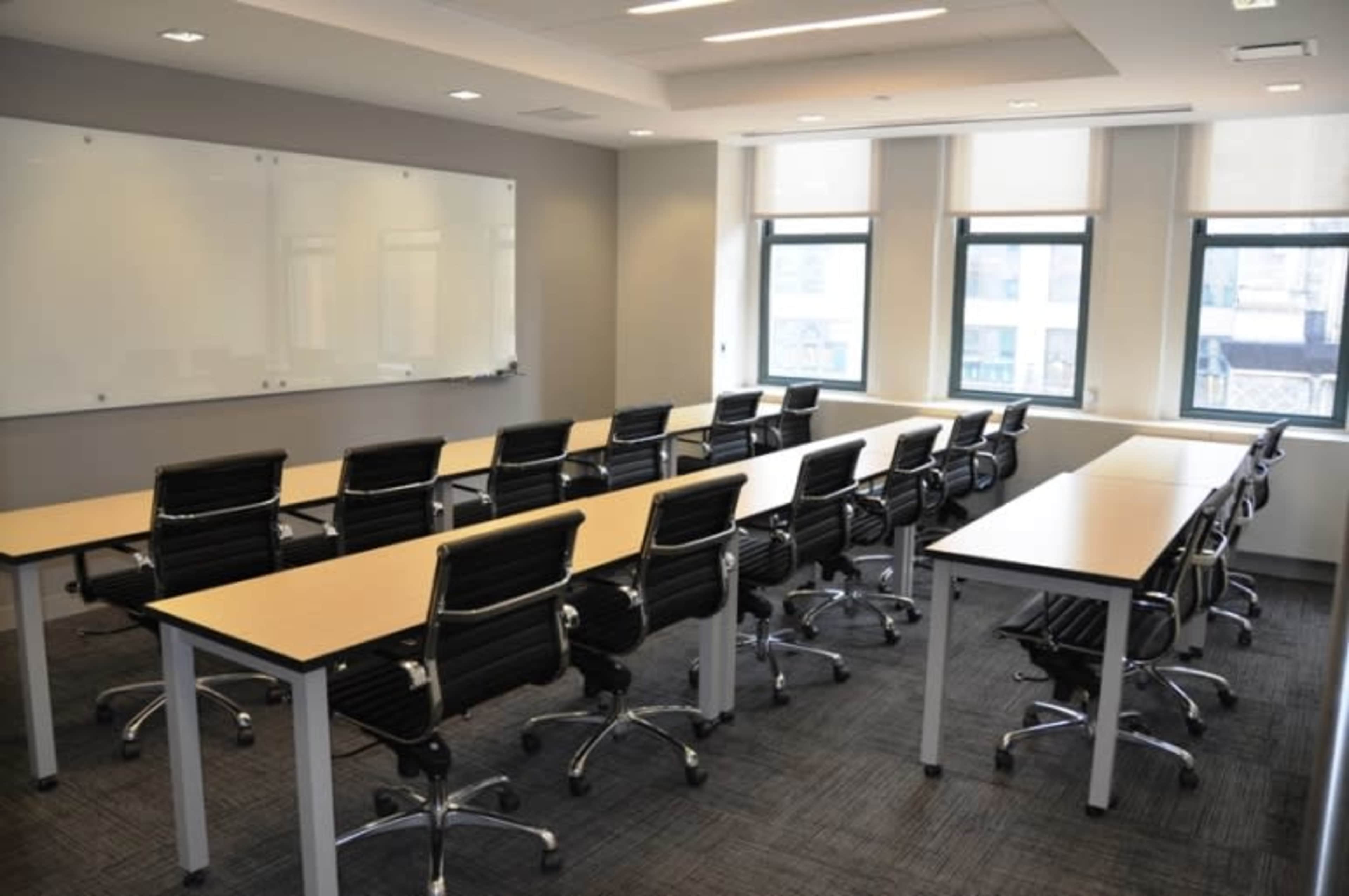 A modern conference room features several rows of desks with rolling chairs, a whiteboard, and large windows.