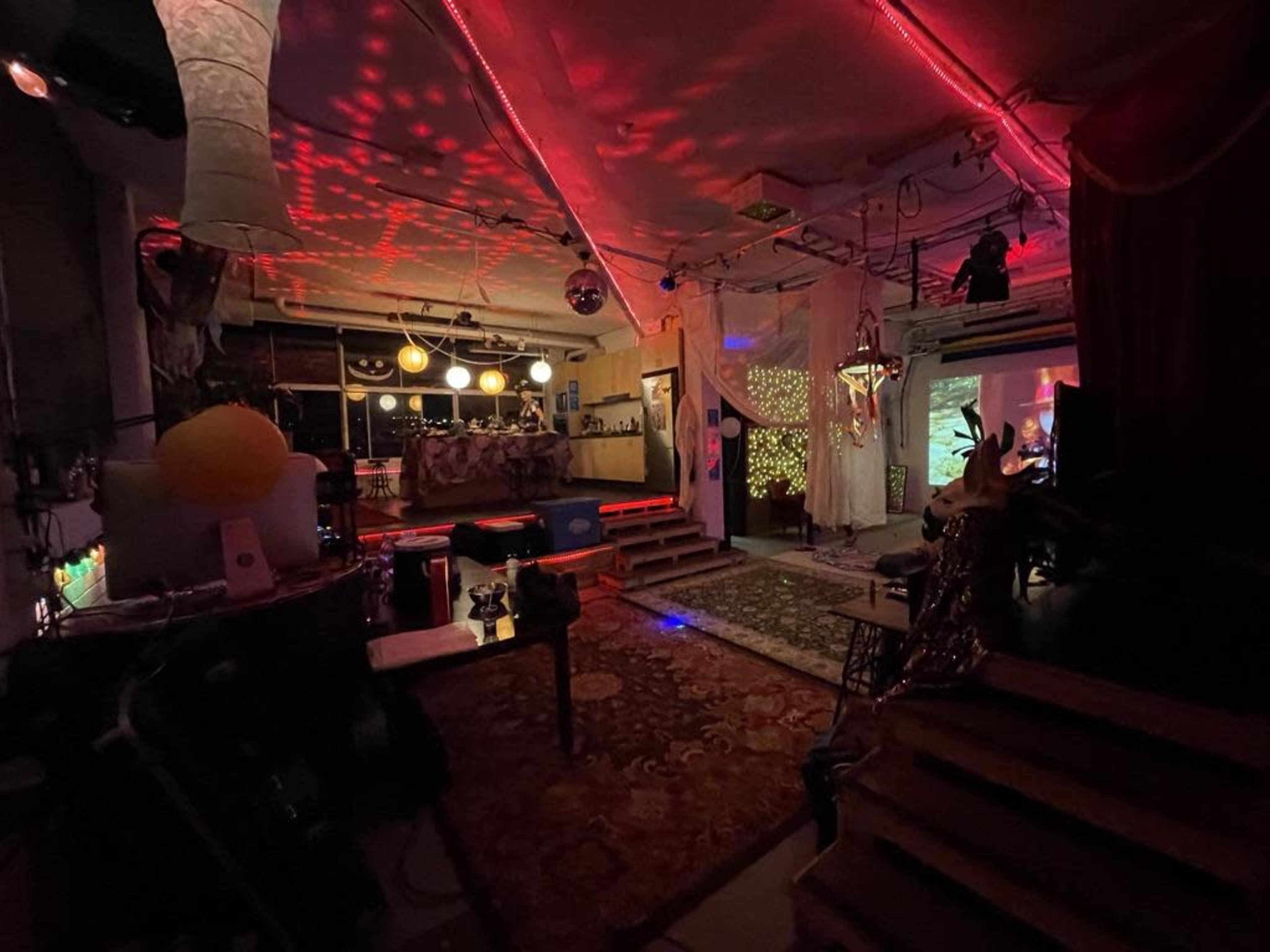 A dimly lit, eclectic interior space with colorful lights, patterned decorations, and various seating arrangements.