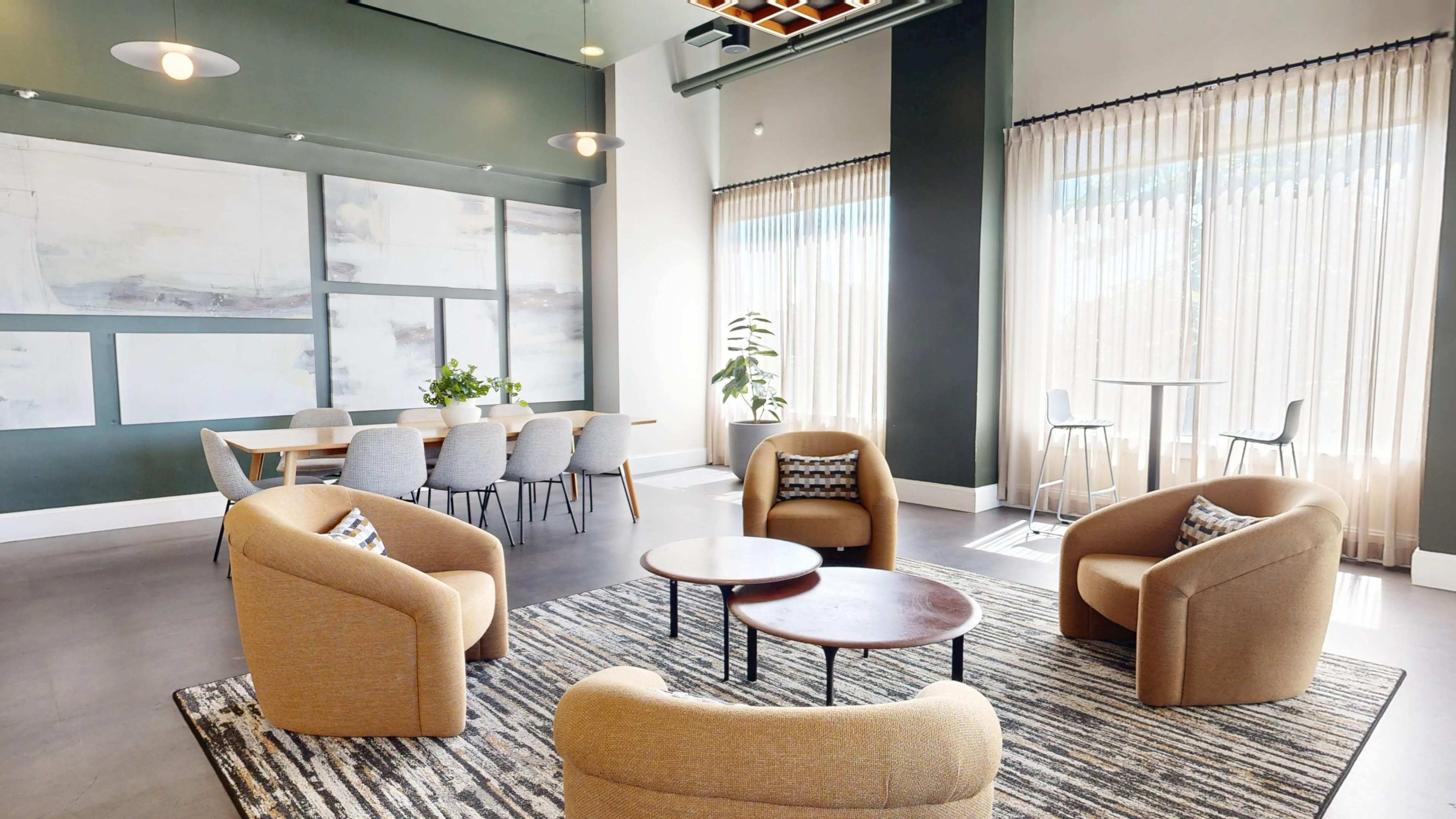 A spacious lounge area features two sets of modern upholstered chairs around low coffee tables, with large windows covered by sheer curtains allowing natural light.