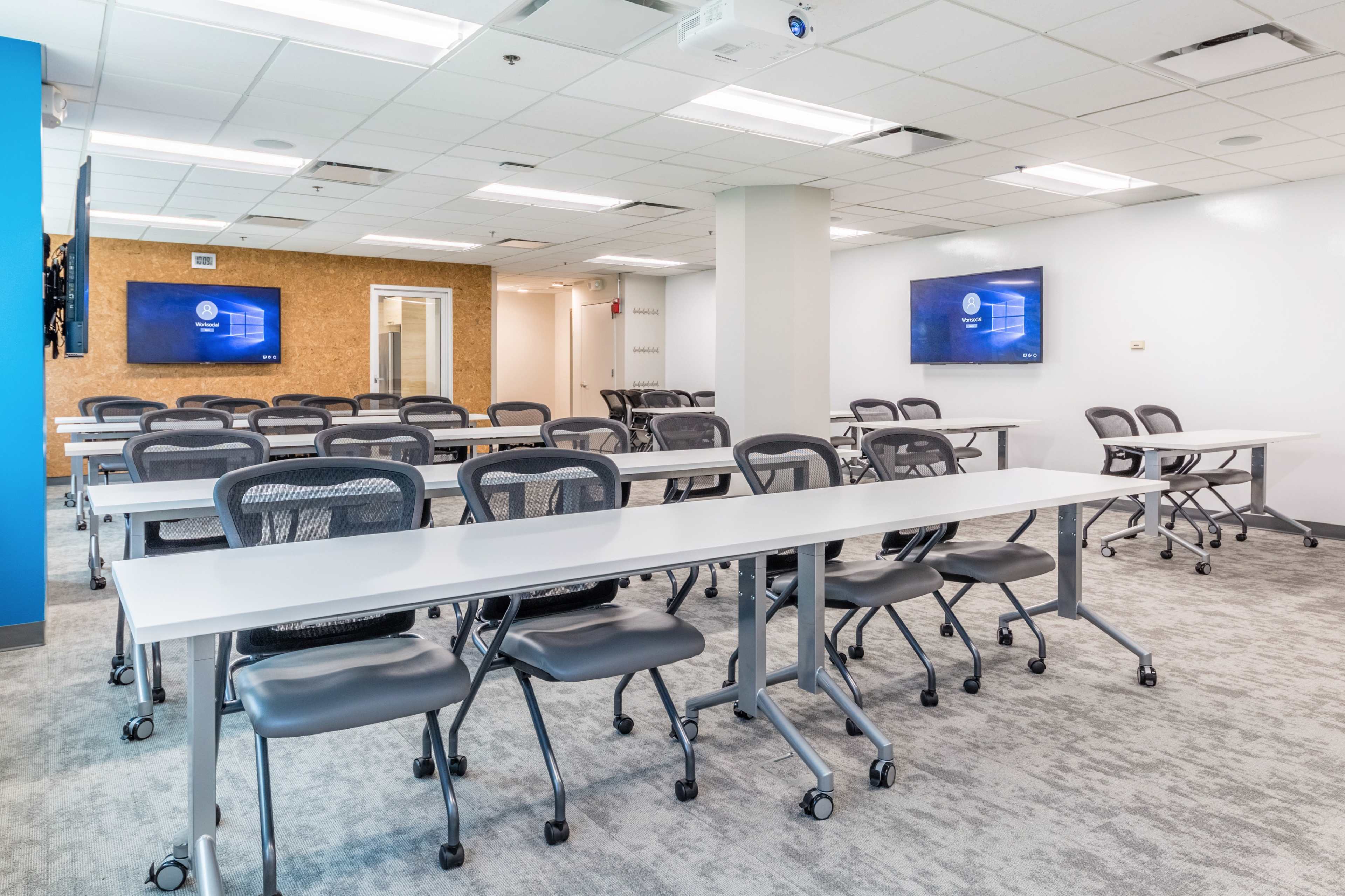 A modern classroom setup with multiple tables and chairs arranged in rows, featuring two wall-mounted screens.