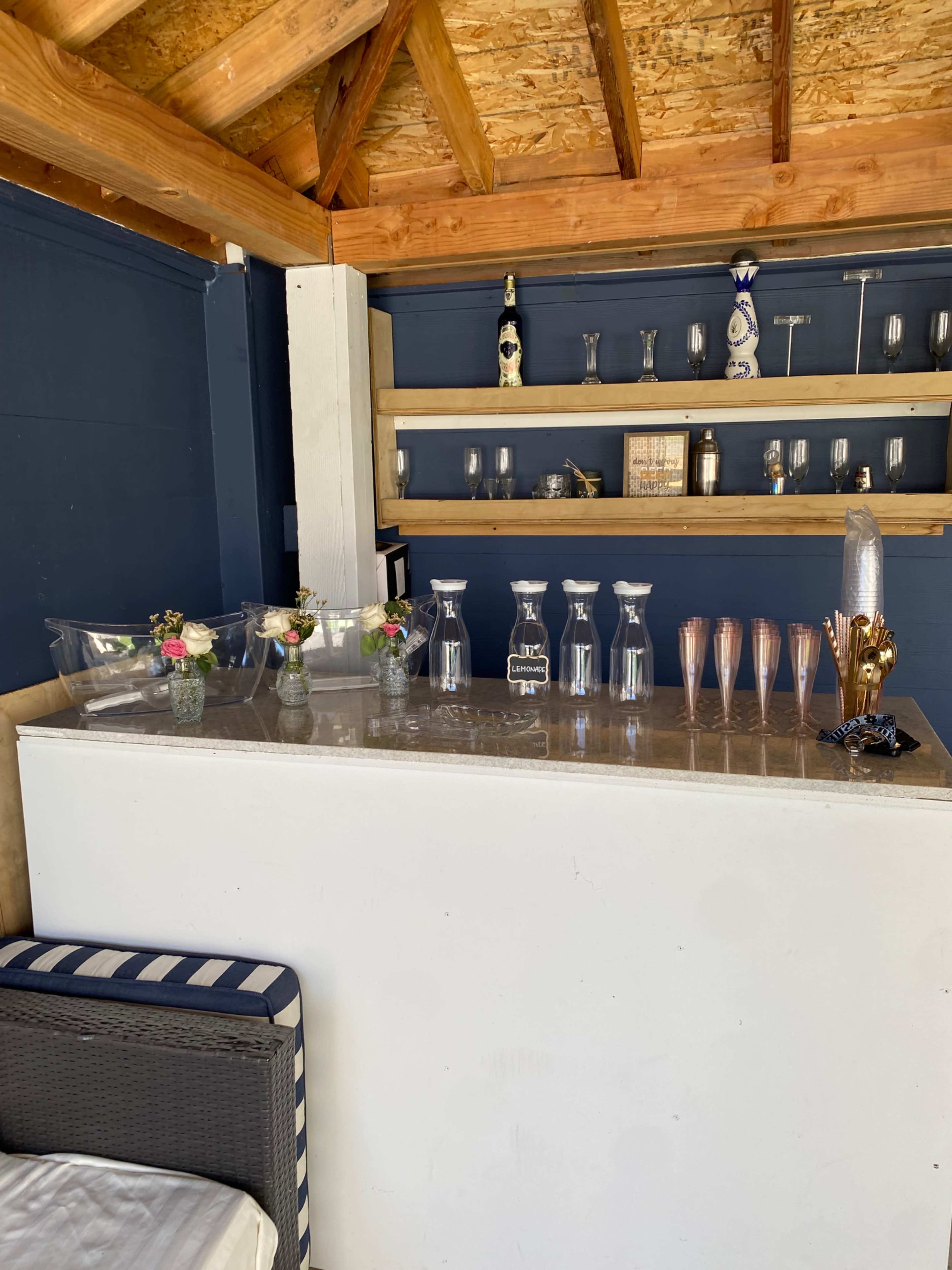 The image shows a bar area with glassware, a silver ice bucket, and decorative flowers on a counter beneath a wooden ceiling.