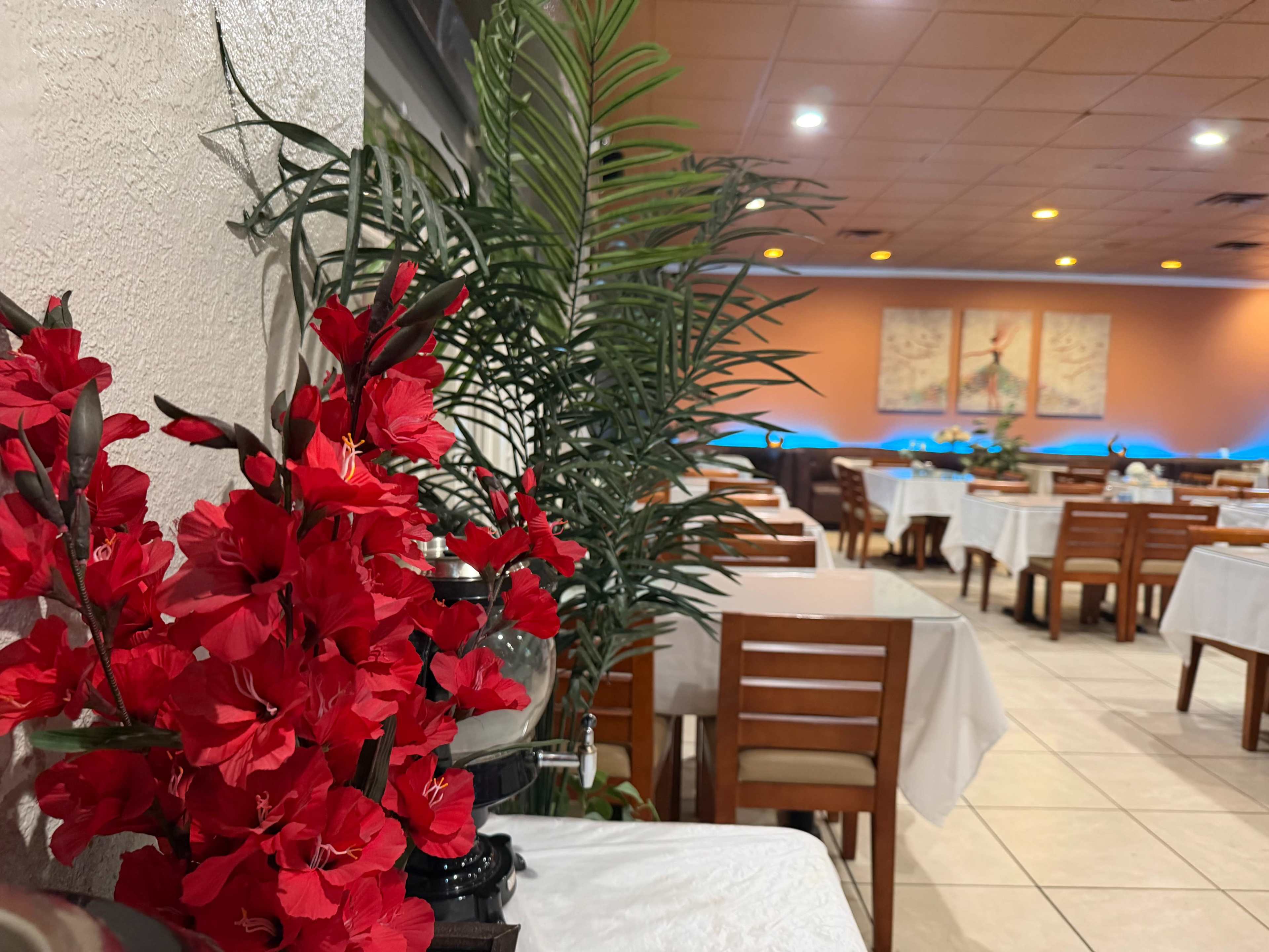 A restaurant interior features tables set with white tablecloths, surrounded by potted plants and red flowers.