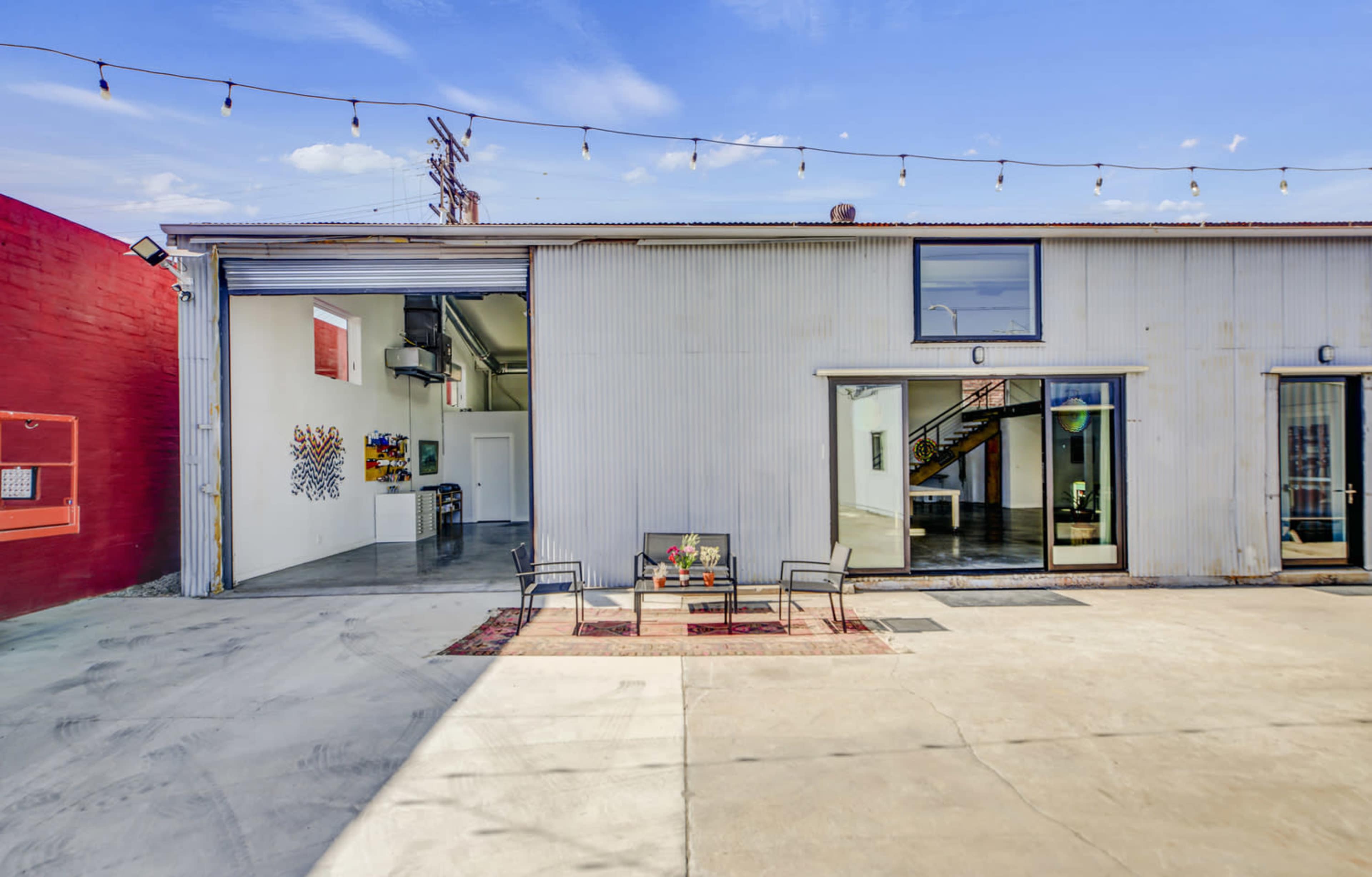 The image shows a spacious outdoor area with a concrete surface, featuring a seating arrangement of two chairs and a small table, alongside a building with large glass doors and a red painted wall in the background.