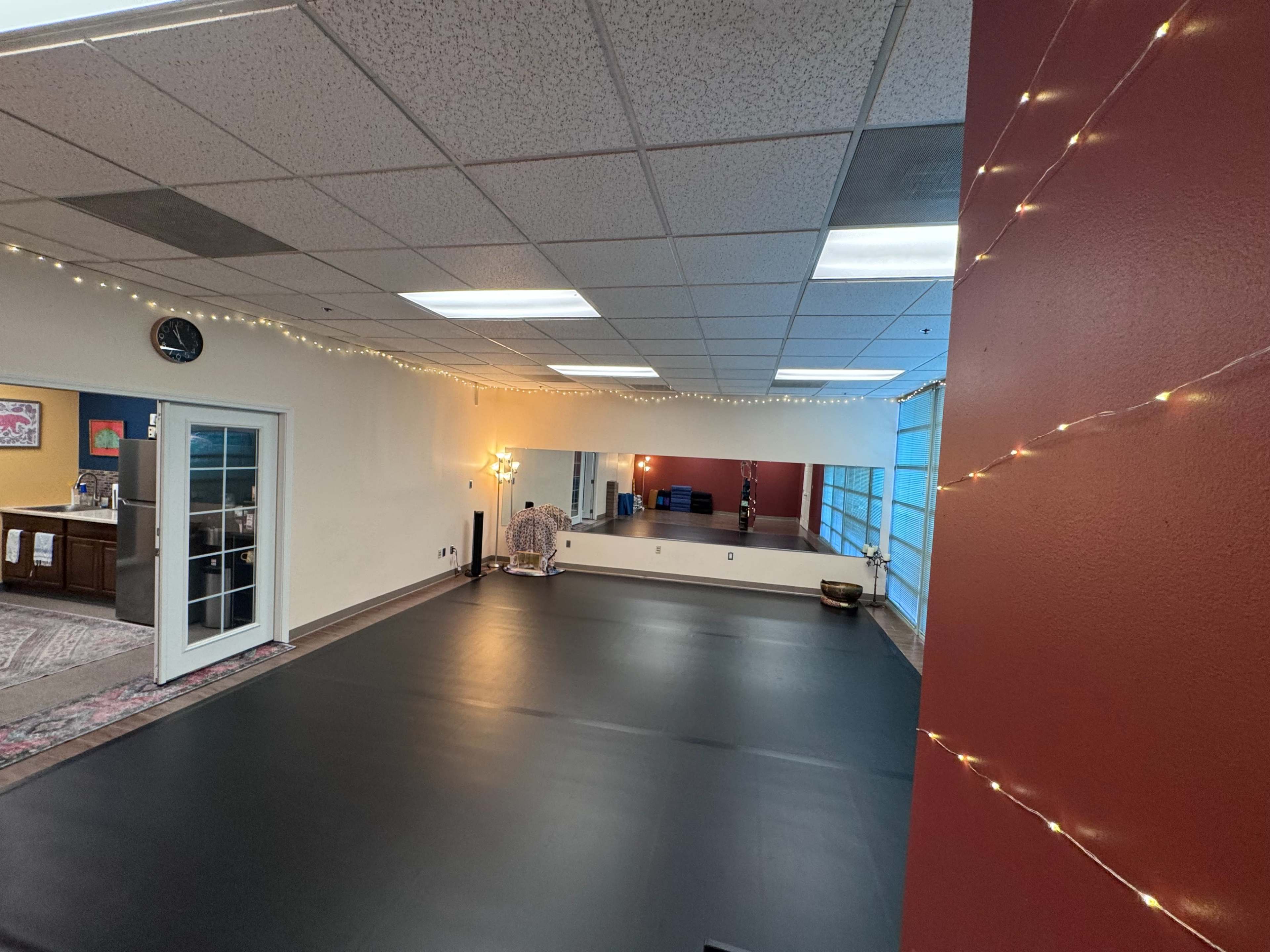 The image shows a spacious, well-lit room with a black mat floor and large windows, decorated with string lights and a seating area.